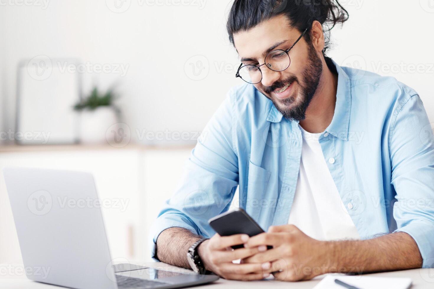 Handsome Eastern freelancer guy sitting at desk at home office and using smartphone, messaging with friends, checking social media or browsing new app, enjoying mobile communication, free space photo