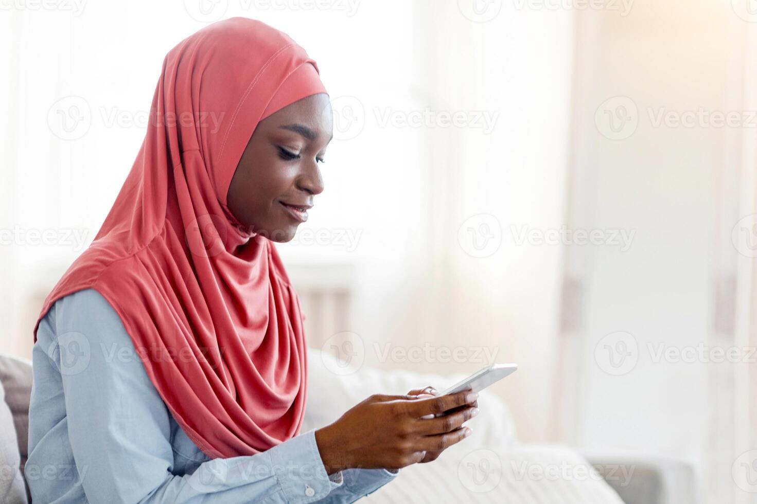 Happy black muslim woman surfing internet on smartphone while resting on couch at home, relaxing in cozy living room with cellphone, sitting on sofa and browsing social networks, side view, copy space photo