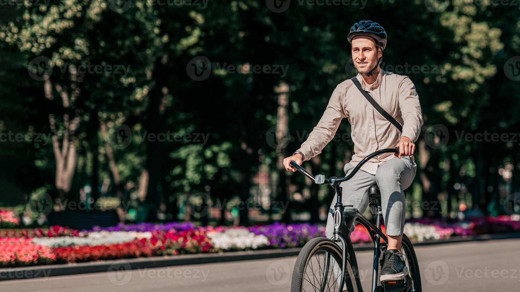 On a bright day, a young man rides his bike down a tree-lined street filled with colorful flowerbeds. He wears casual attire and a helmet, embracing an active lifestyle in the city, copy space photo