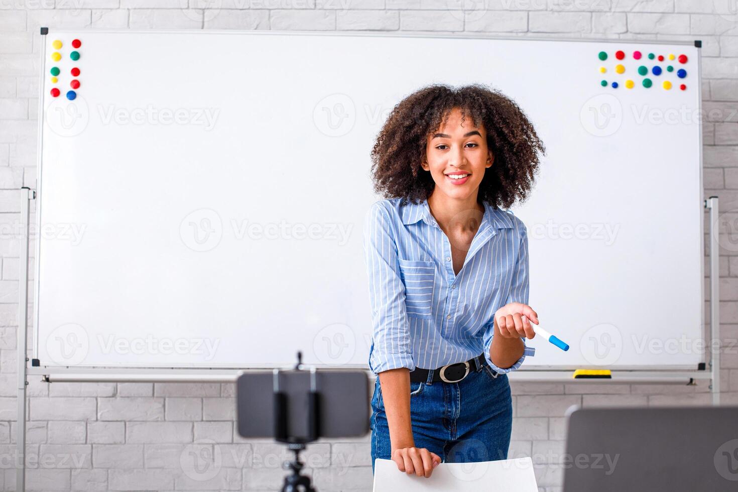 Free time hobby or work remotely during covid quarantine. Smiling african american young woman explaining rules of english and looking at smartphone camera at table with laptop in living room photo