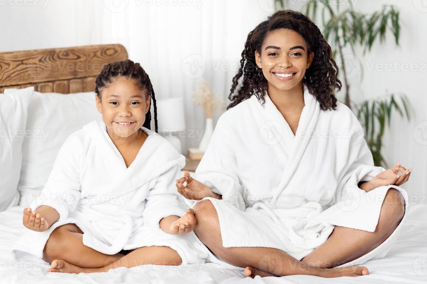 Wellness Day. Cheerful African American Mom And Daughter In Bathrobes Sitting In Lotus Position, Practicing Yoga Together After Bath, Meditating On Bed In Cozy Room, Smiling At Camera, Copy Space photo