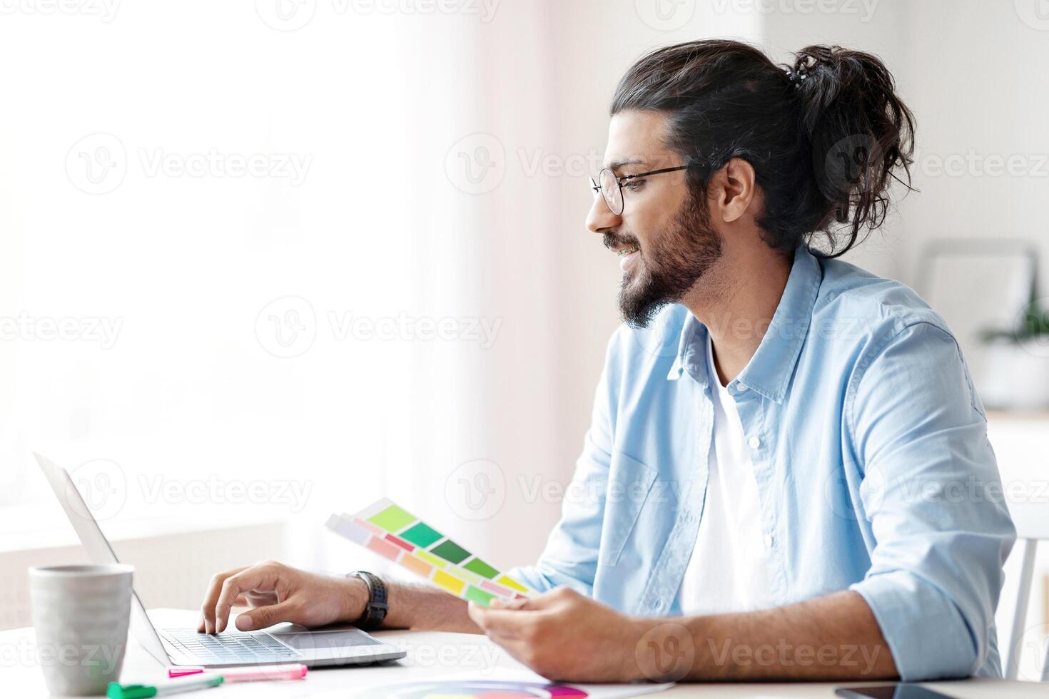 Handsome young eastern man interior designer holding color swatch samples and working on laptop at home office, checking palette for new design project, sitting at desk near window, copy space photo