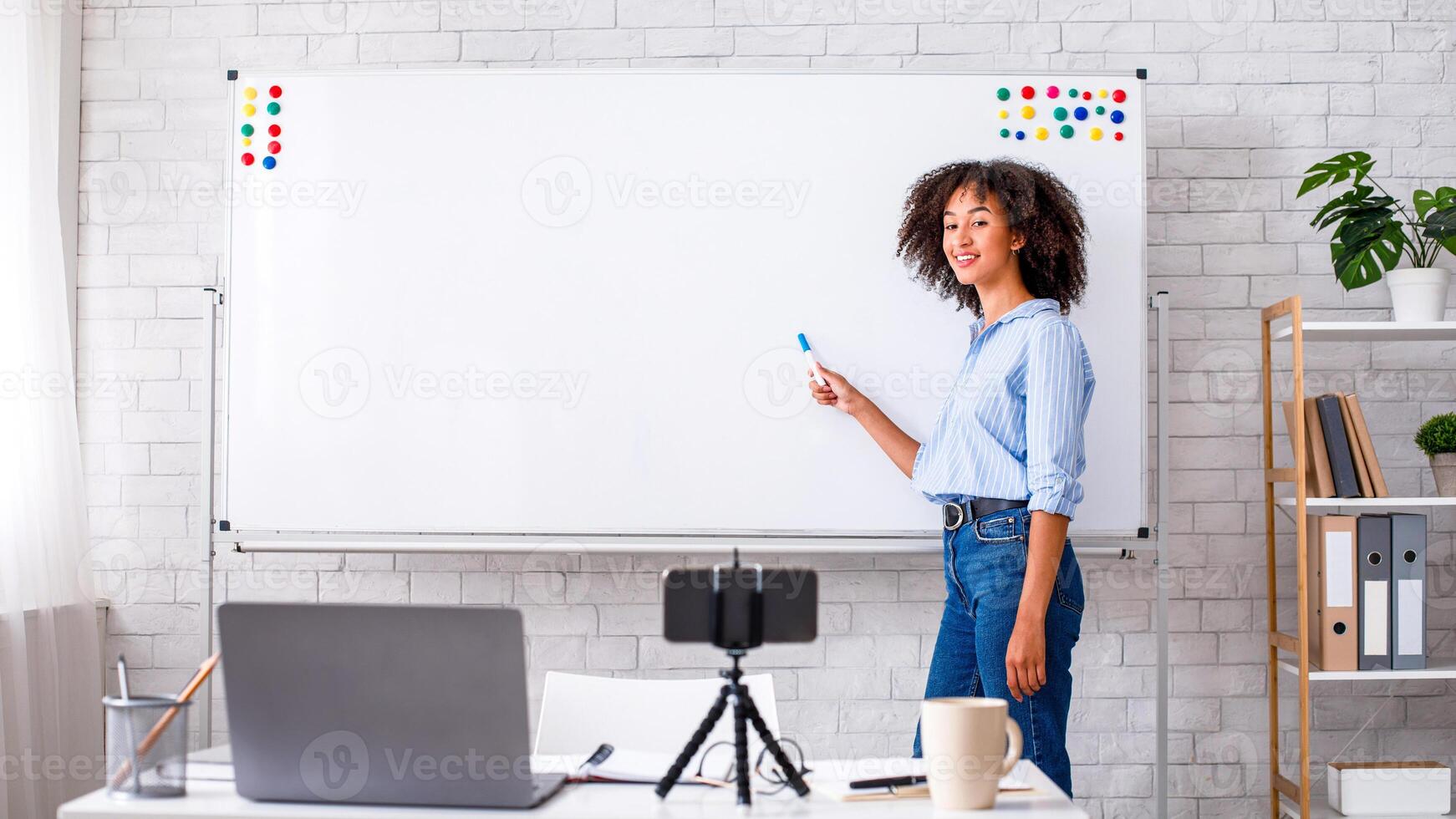 Online education and English lesson. Friendly african american woman points to blackboard with rules and watching in webcam of smartphone on desk with laptop in home class interior, copy space photo