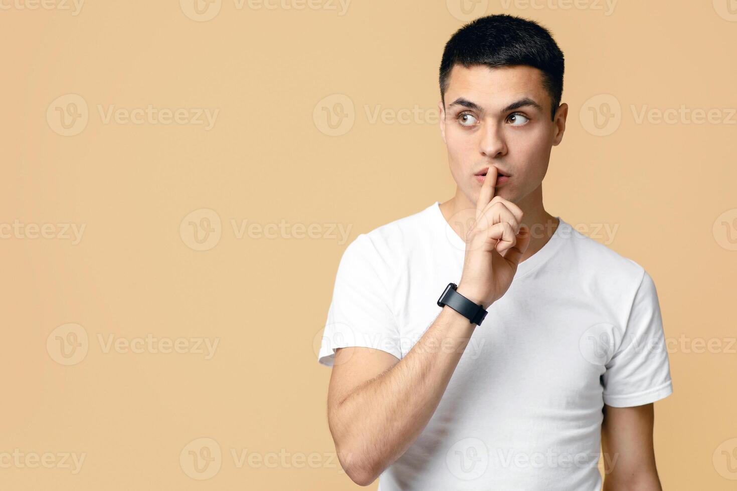 Keep secret, be silent, great offer and advertising. Serious young blogger student man in white t-shirt with smart watch makes secret sign, isolated on light sand background, empty space, studio shot photo