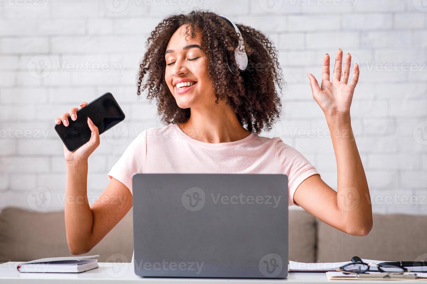 Take a break from work at home. Happy african american woman with headphones singing at smartphone at table with laptop in living room interior, free space photo