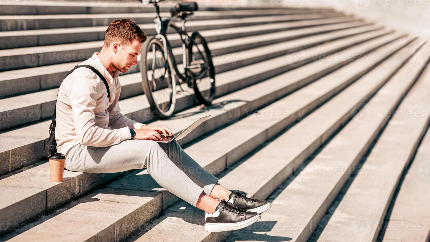 Urgent work and deadline in city outdoors. Busy millennial man manager freelancer or blogger sits on stairs in street, works on laptop, with takeaway coffee and bike nearby photo