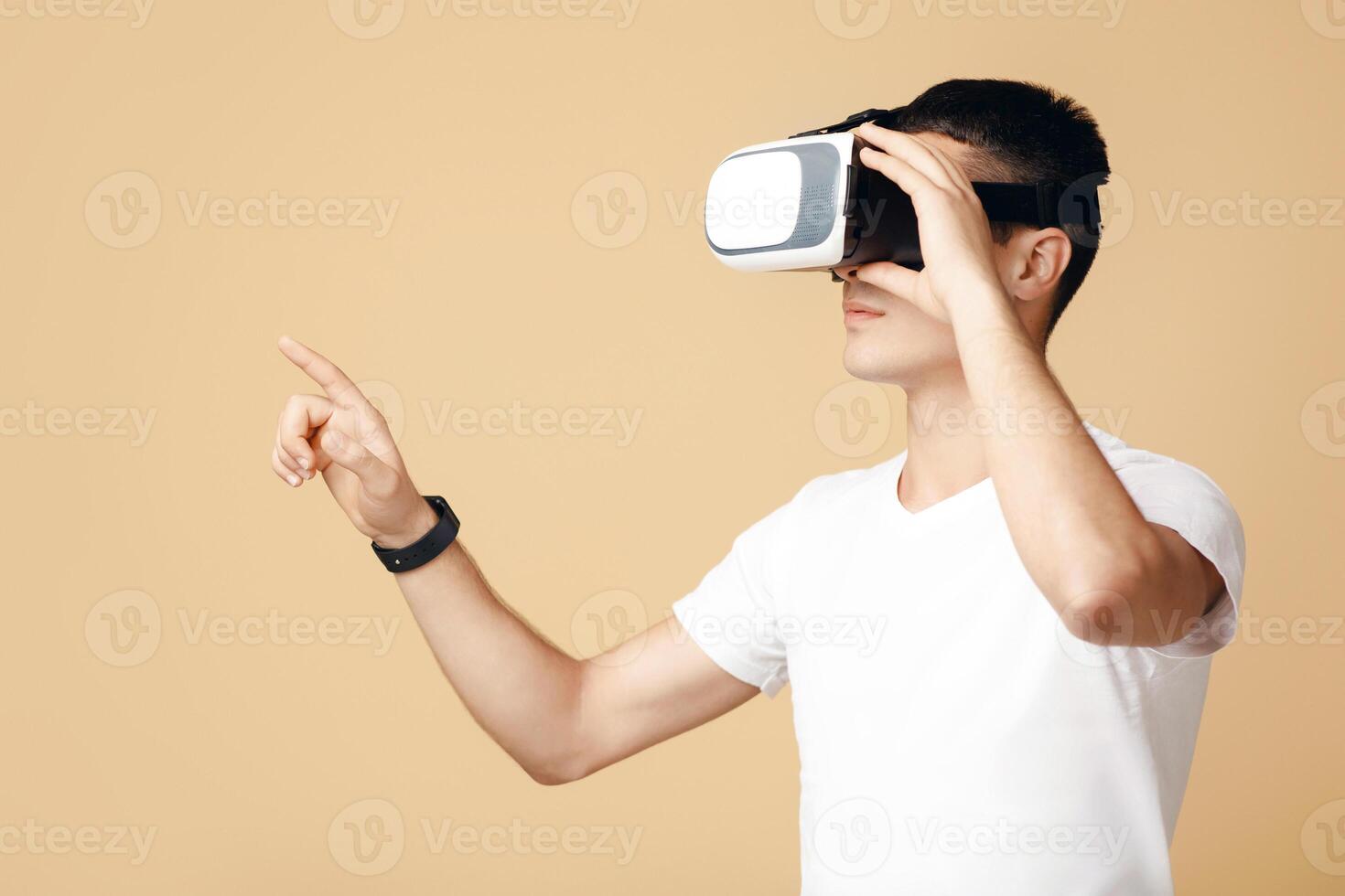 Man in goggles playing in games. Young student in white t-shirt and virtual reality spectacle touches to blank space with finger, isolated on sand or light background, side view, studio shot photo