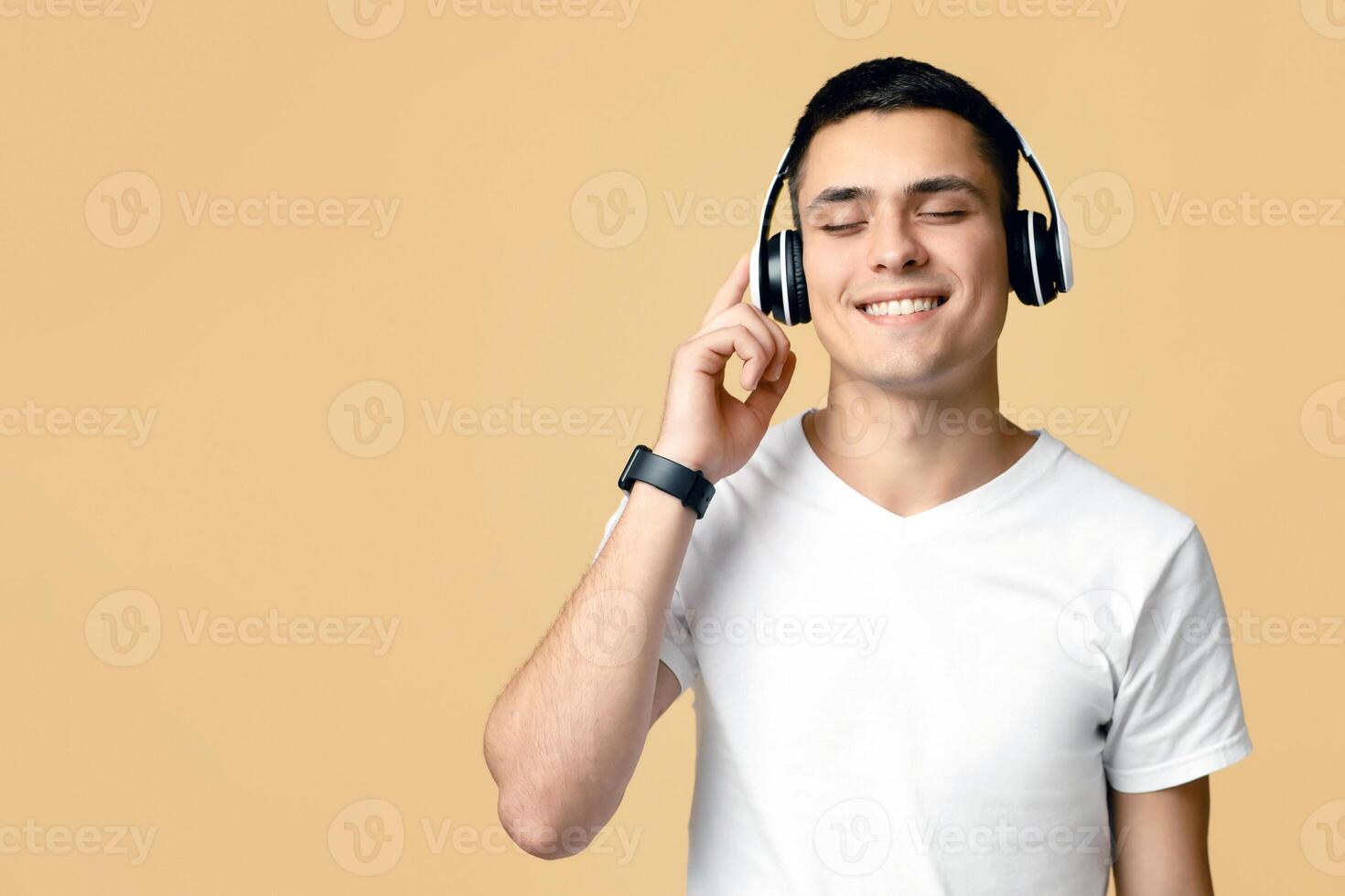 Enjoy music, stay at home. Smiling young blogger student man in white t-shirt with smart watch listens to song in modern headphones, isolated on light background photo