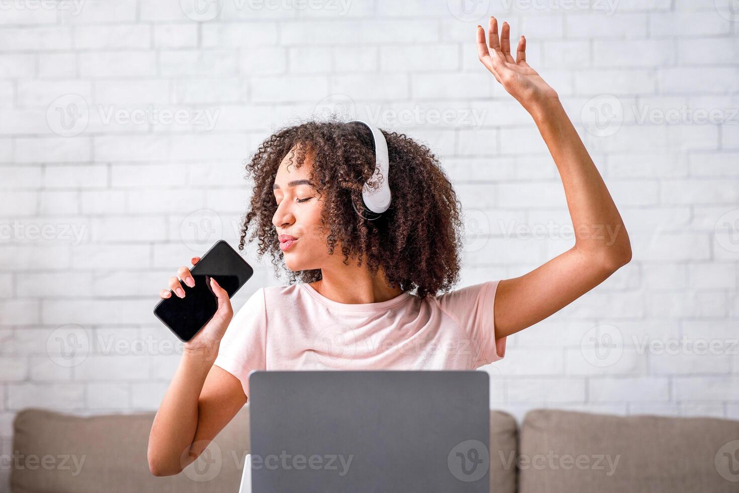 Fun music break and vocals app. Cute african american woman in headphones with closed eyes enjoys and sings at smartphone with empty screen, sitting at table with laptop in living room interior photo