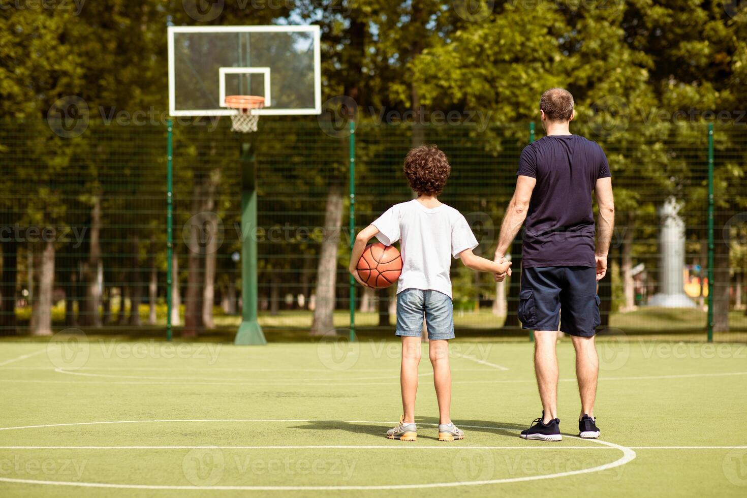 Healthy Lifestyle. Rear view of dad and son standing on playground with basket ball, holding hands, looking at hoop photo