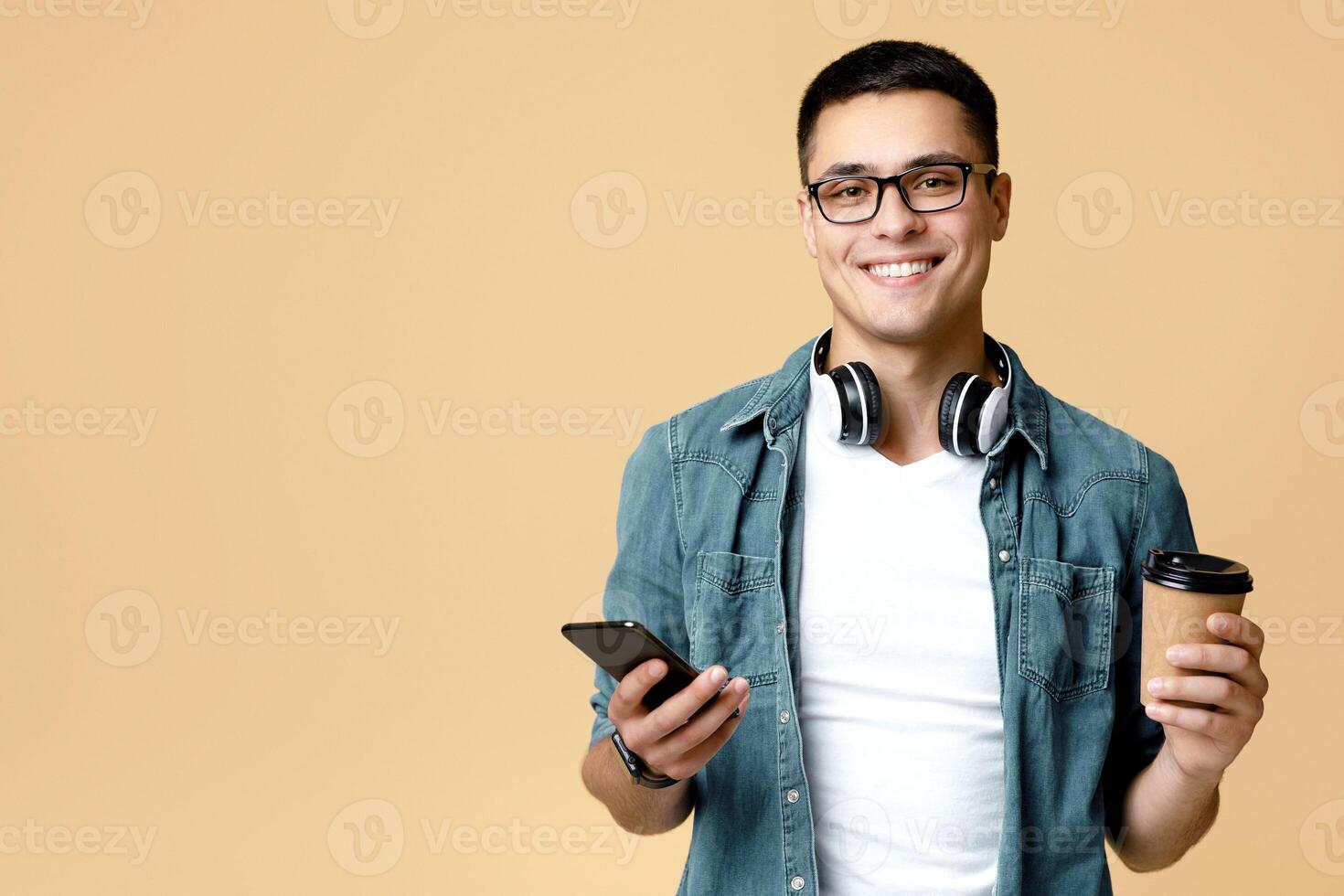 Modern hipster, employee or blogger and positive emotions. Smiling young man student in white t-shirt and glasses holds phone and cup of coffee to go, isolated on beige background, studio shot photo