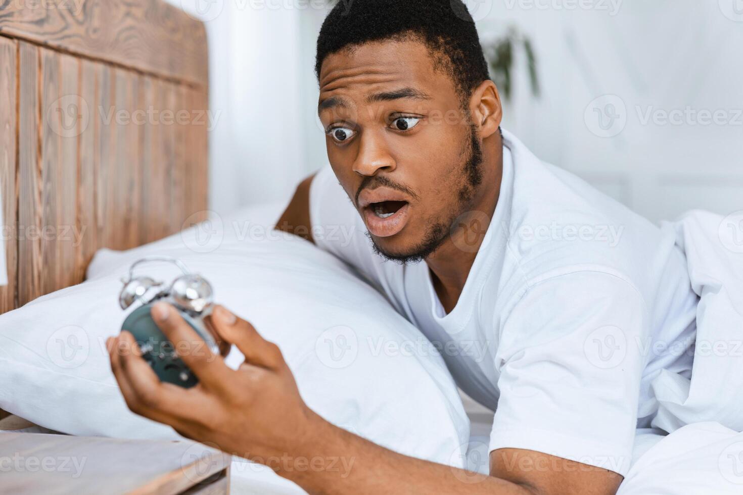 Oversleeping. Overslept African American Man Looking In Shock At Broken Alarm Clock Waking Up In The Morning, Lying In Bed At Home. Late For Work, Bad Morning, Tardiness Concept photo