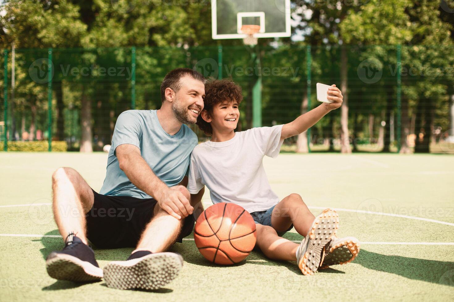 Portrait of happy dad and son taking selfie using smart phone, sitting on sports ground, selective focus photo