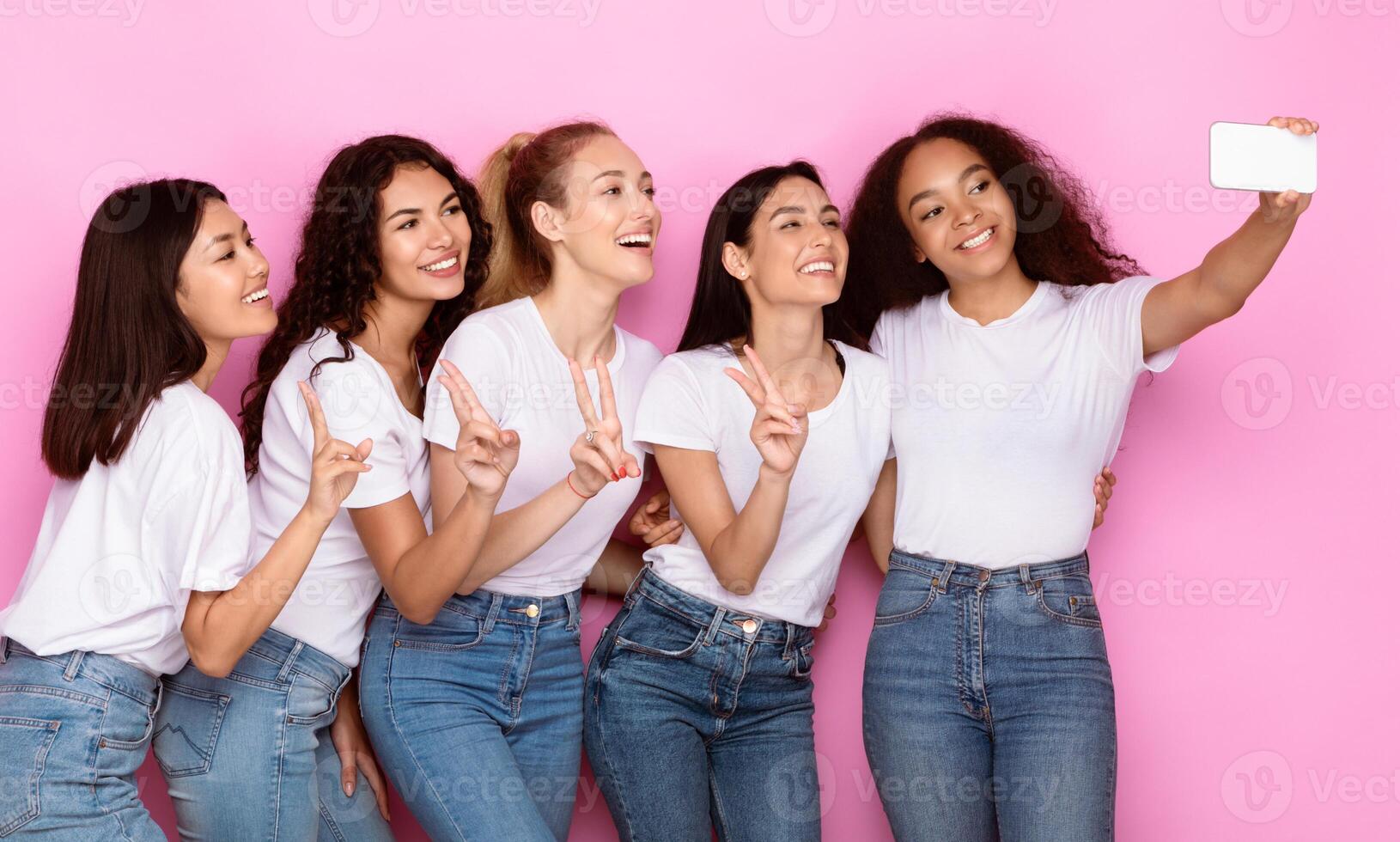 Diverse Female Group Selfie. Cheerful Multiracial Women With Smartphone Having Fun Making Their Self-Portrait On Phone Posing On Pink Studio Background. Female Friendship Concept. photo
