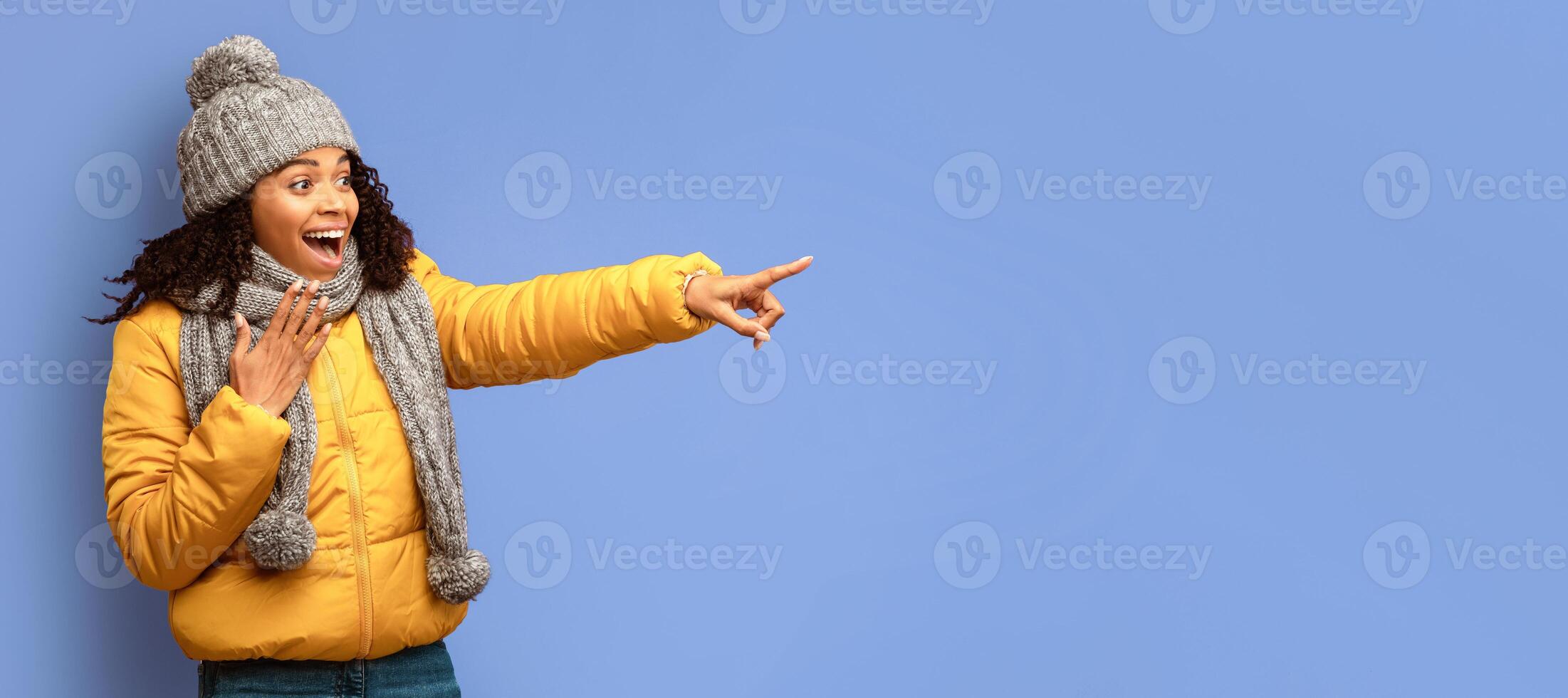 Look There. Excited Black Woman In Winter Jacket Pointing Finger Aside At Copy Space Posing On Purple Studio Background. Panorama photo