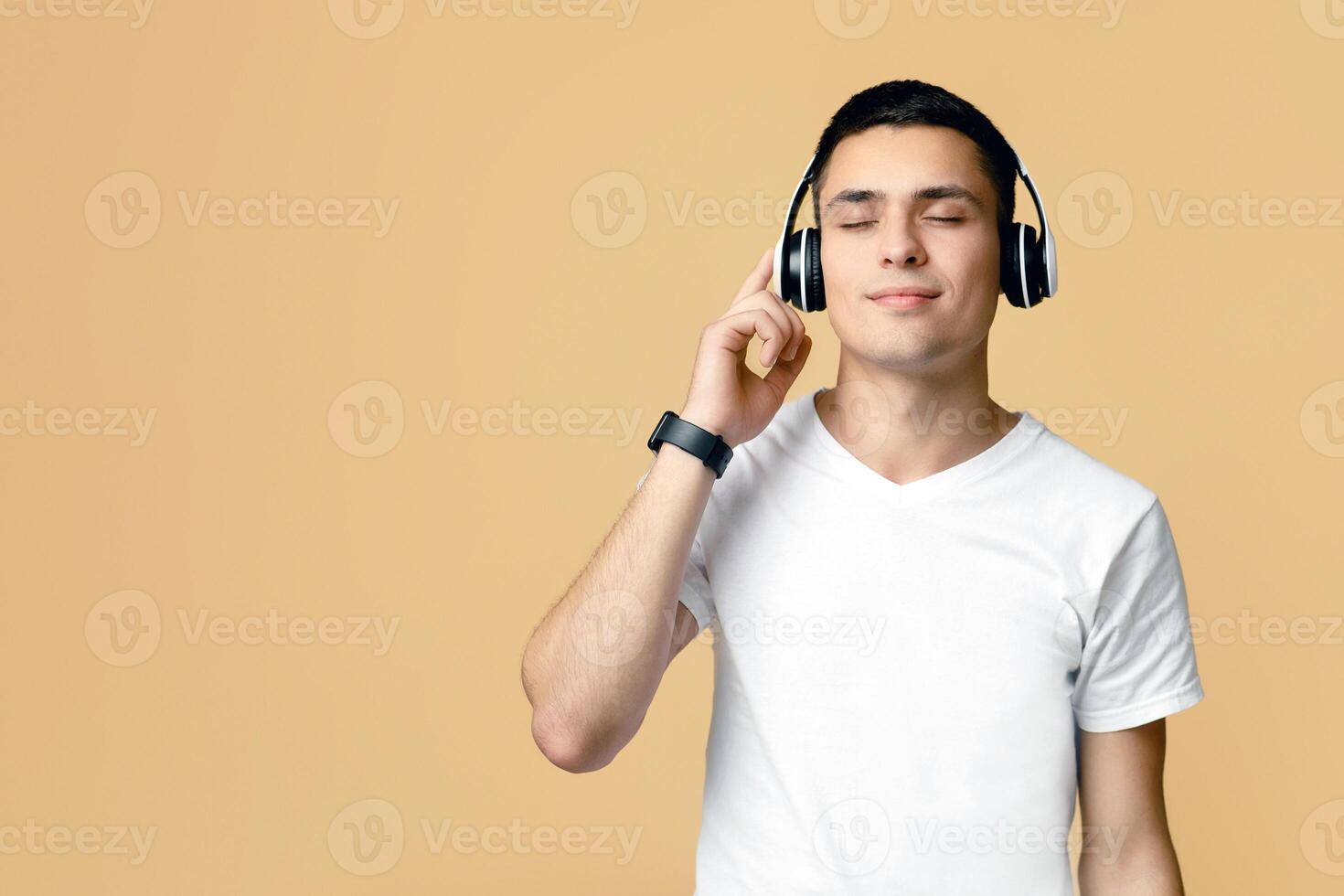 Music fan, favorite songs, break and relax. Peaceful young man student with closed eyes in white t-shirt and headphones listening music, isolated on beige background, studio shot, free space photo