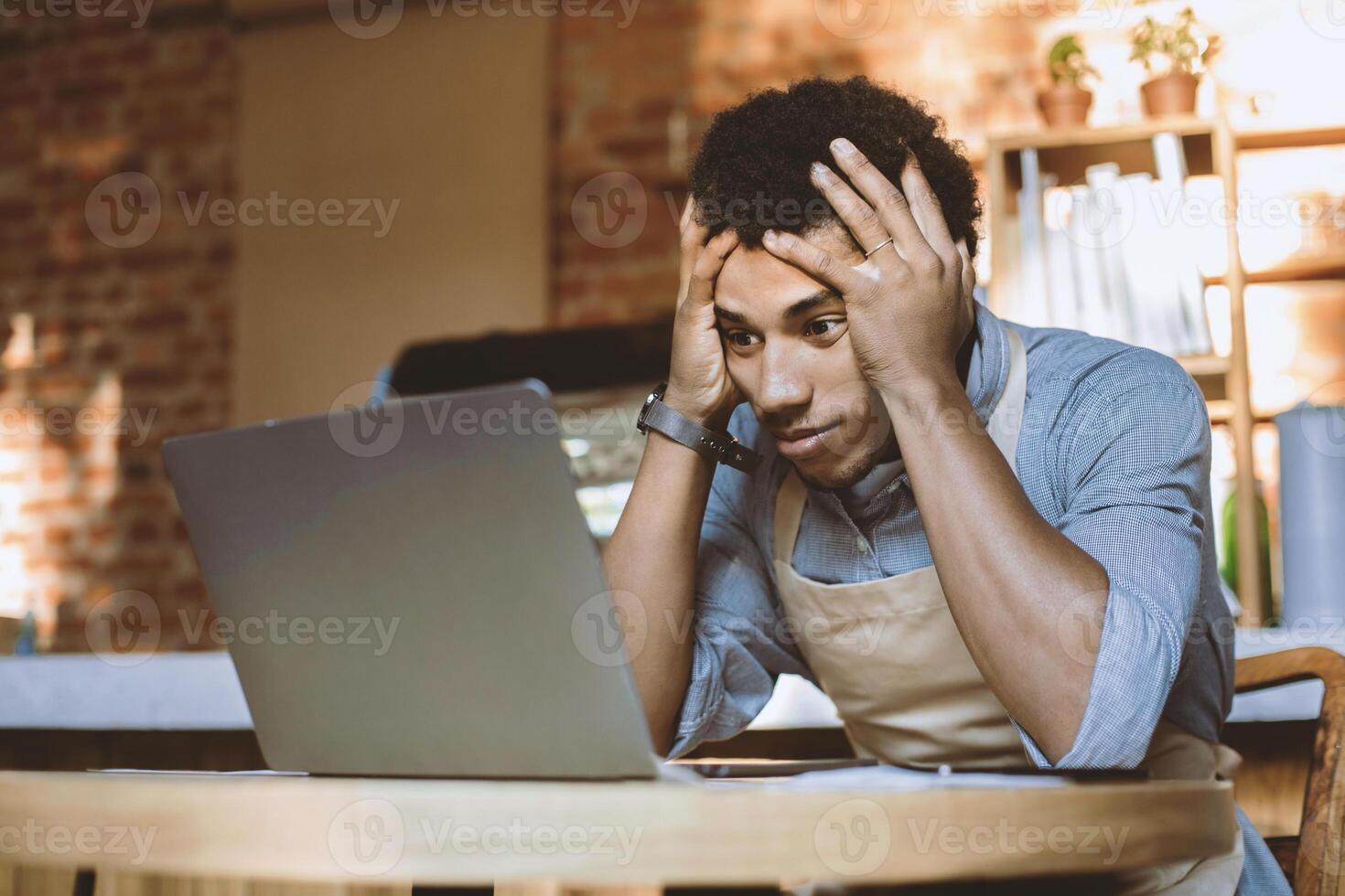 Ruin and financial problems during covid-19 quarantine. Sad young african american guy in apron holds head and looks in laptop at financial accounts and big debt in modern cafe, empty space photo