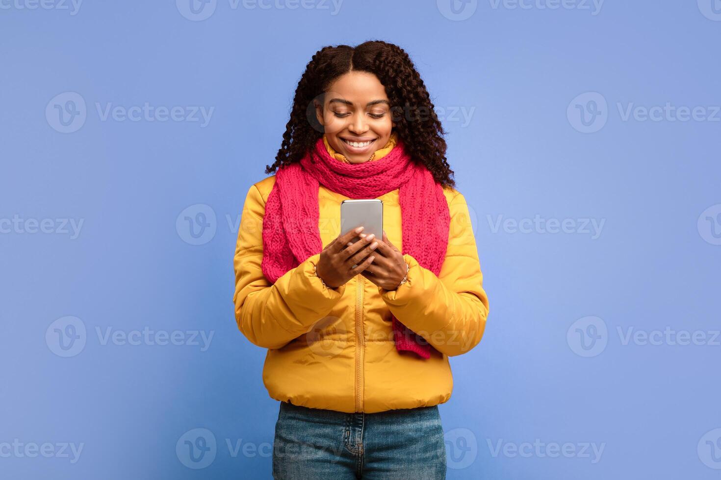 African American Woman Using Smartphone Phone Networking Texting In Social Media Mobile App Standing Over Purple Studio Background, Wearing Winter Clothes. Cellphone Application Concept photo