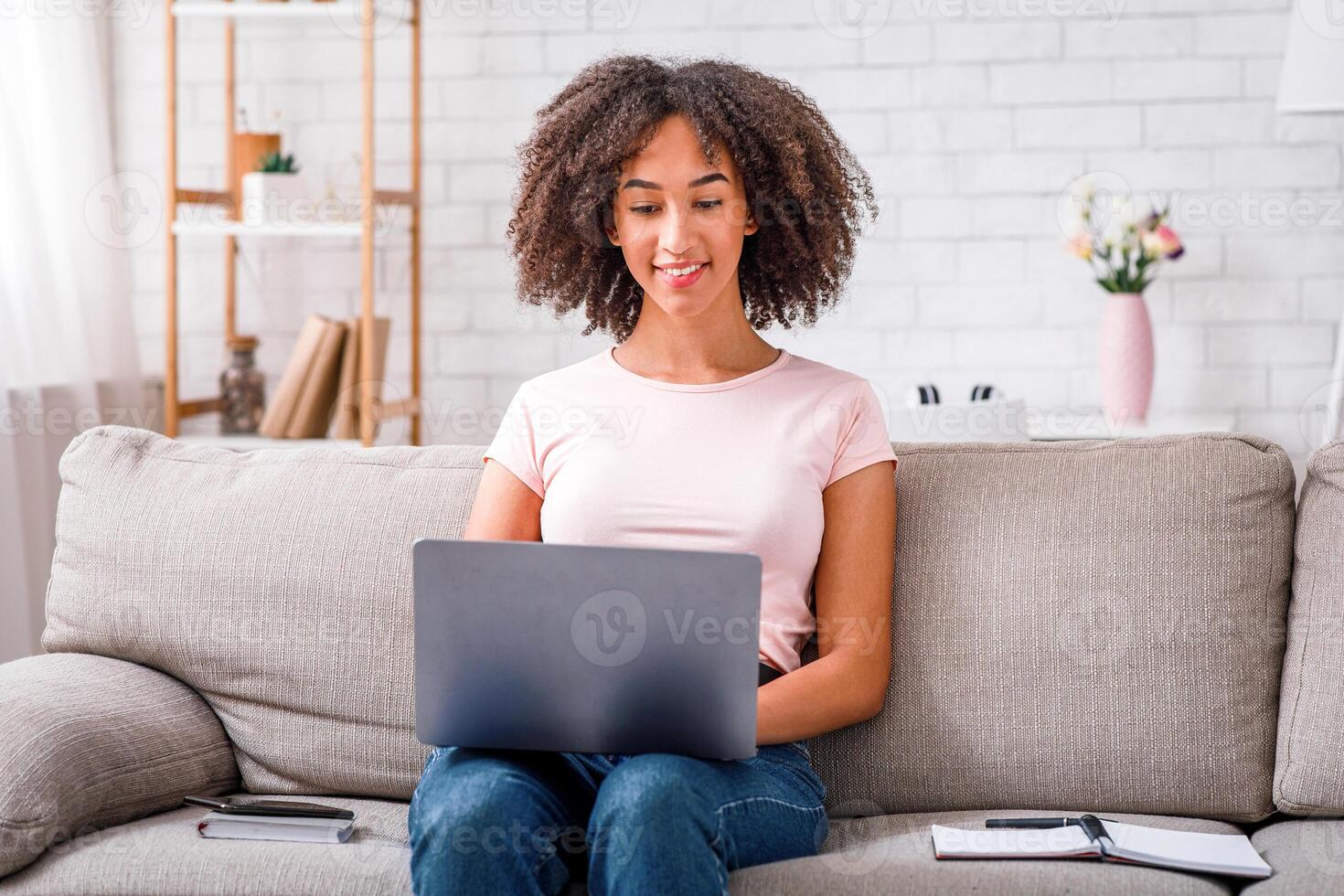 Online work in social networks during quarantine and self-isolation. Friendly african american girl working in laptop, sitting on couch with notepad photo