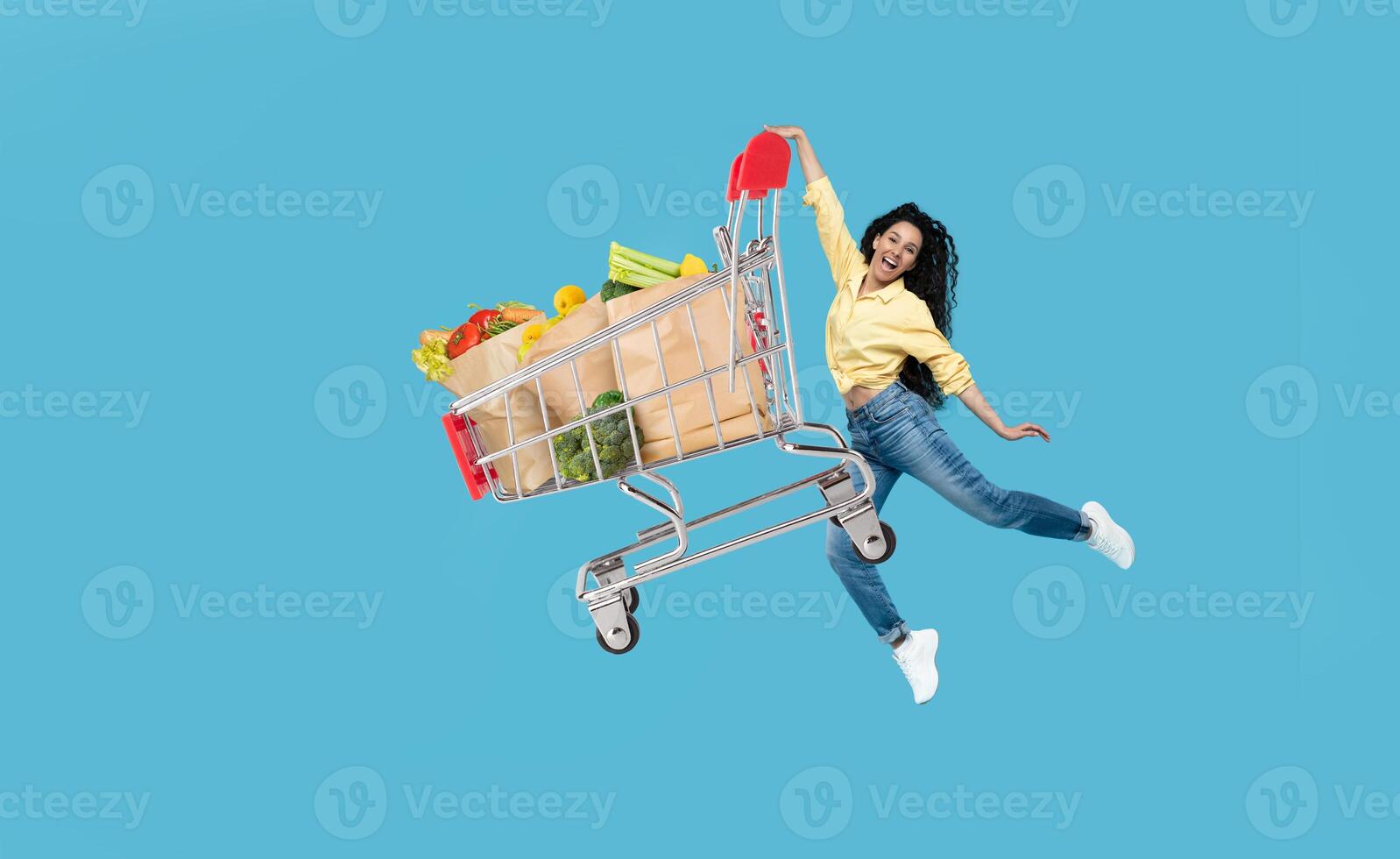 A joyful shopper is seen exuberantly pushing a grocery cart filled with fresh produce and items against a vibrant blue background. This captures the spirit of modern purchasing. photo