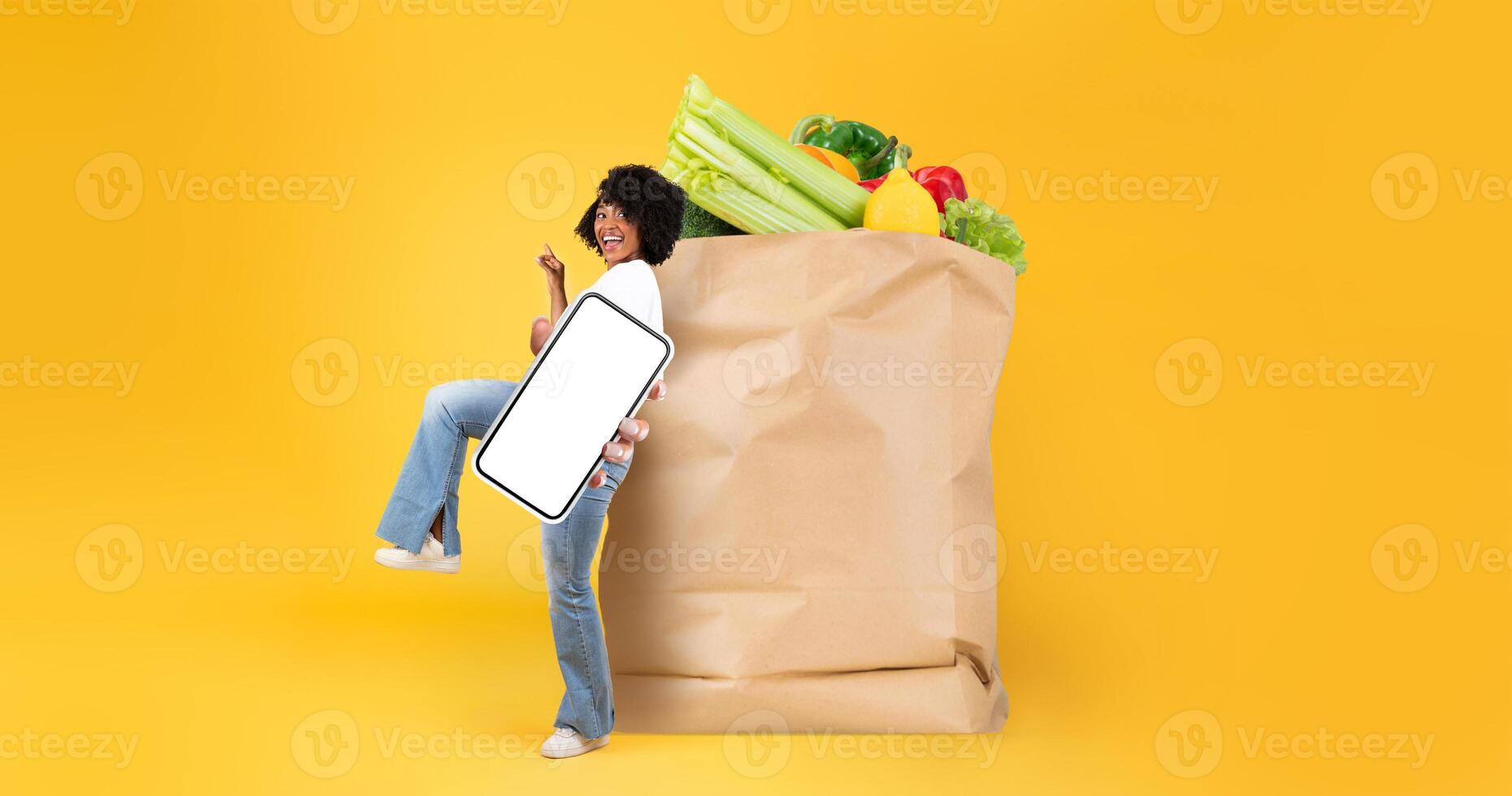 A customer happily engages in grocery shopping, holding a mobile phone in one hand while carrying a large paper bag filled with fresh produce. photo
