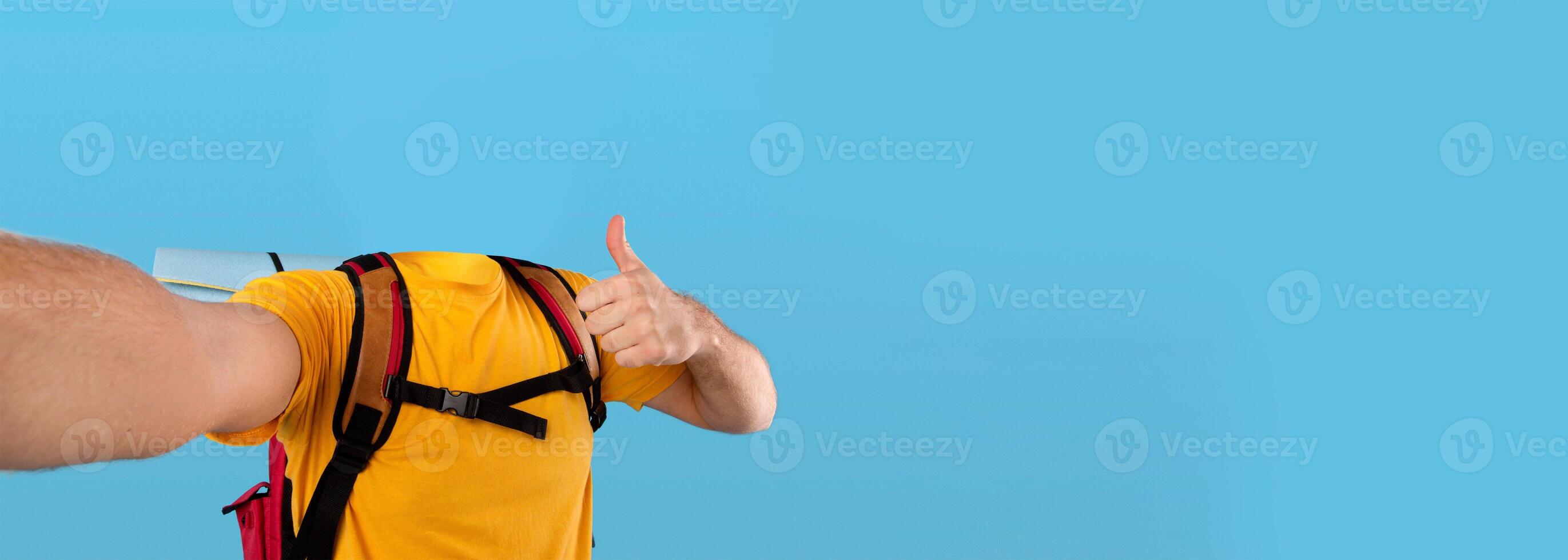 A headless individual in a yellow shirt and backpack is making a thumbs up gesture, taking selfie. The isolated setting with a blue background emphasizes a positive travel vibe and adventure spirit. photo