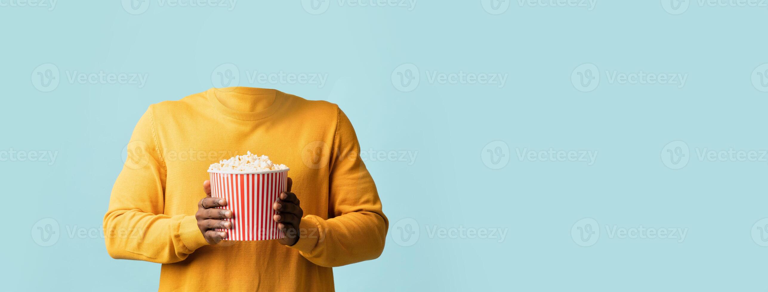A headless figure dressed in a yellow sweater holds a large striped popcorn bucket against a soft blue background. This isolated composition emphasizes lifestyle themes and fun activities. photo