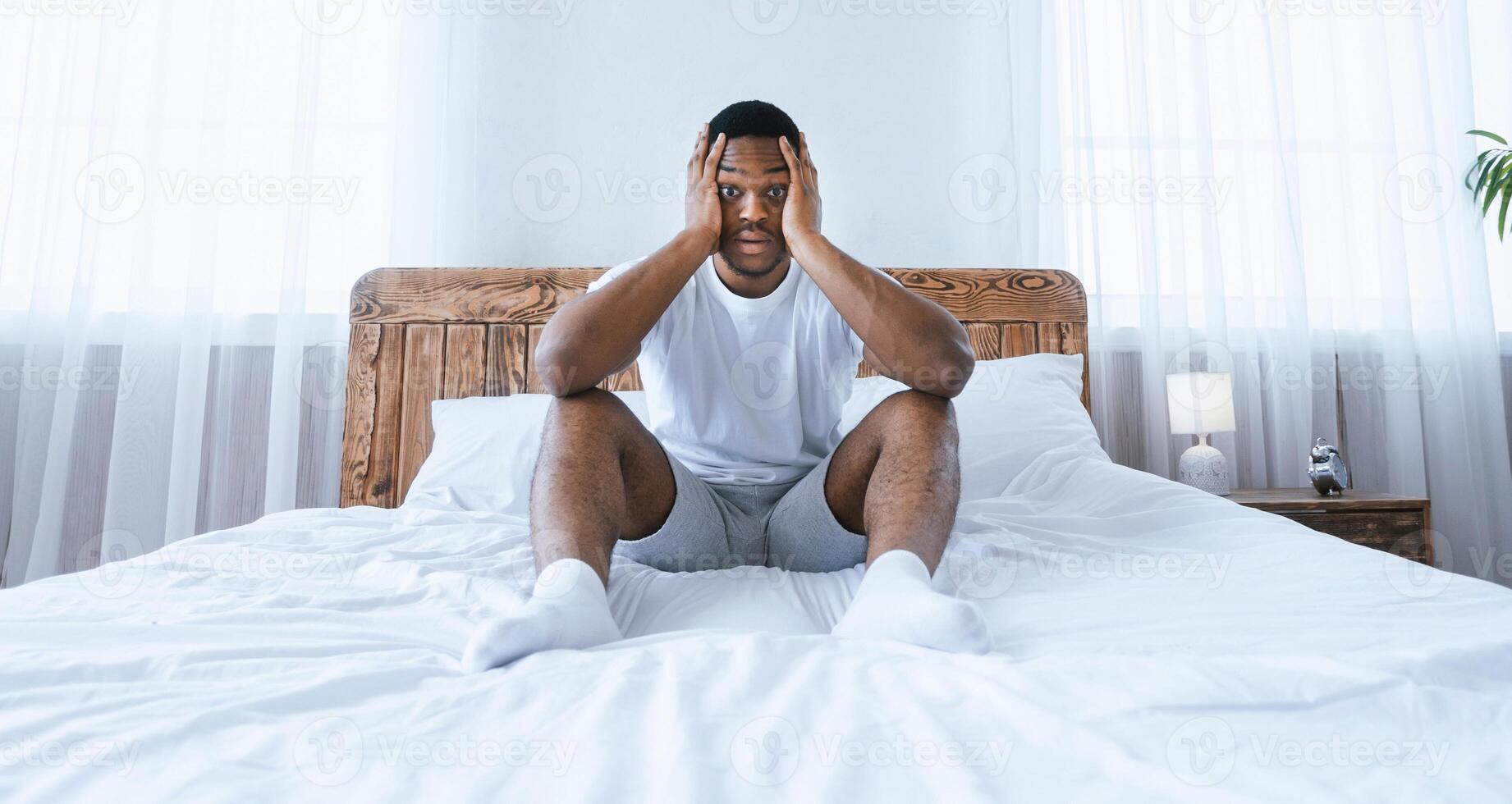 Insomnia And Oversleeping. Stressed Overslept African American Man Sitting In Bed, Touching Head Looking At Camera In Shock In Bedroom At Home. Stressful Life, Sleeplessness Concept. Panorama photo