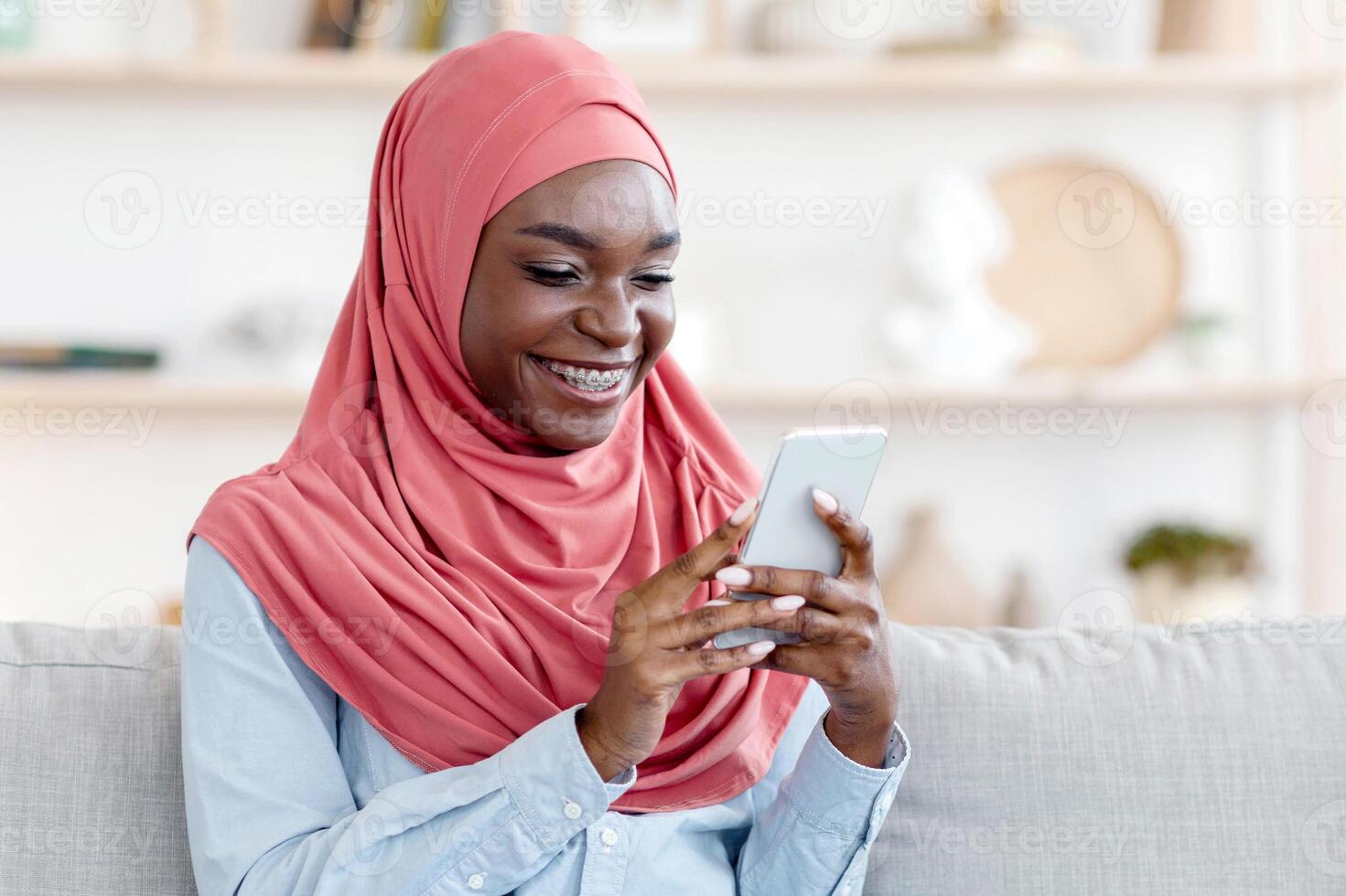 Social Networks. Young black muslim female in hijab using smartphone at home, sitting on cozy couch, messaging with friends, browsing internet, relaxing in living room, enjoying weekend, copy space photo