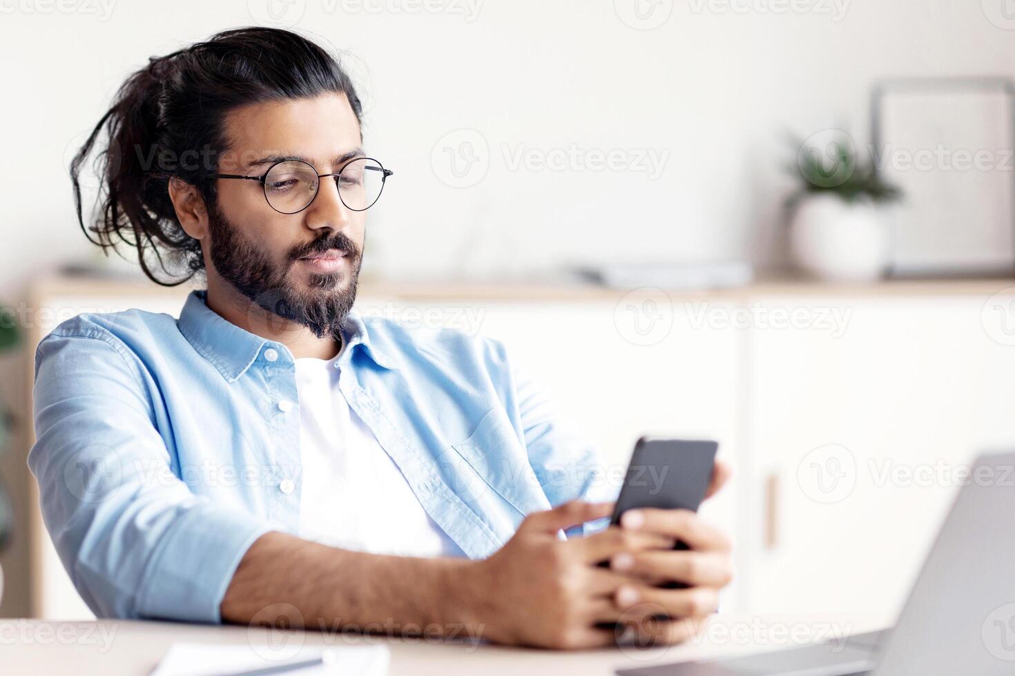 Young Arab Male Entrepreneur Relaxing At Workplace With Smartphone, Browsing Internet Or Texting With Friends, Handsome Eastern Manager Guy Sitting At Desk With Laptop, Having Break In Work photo
