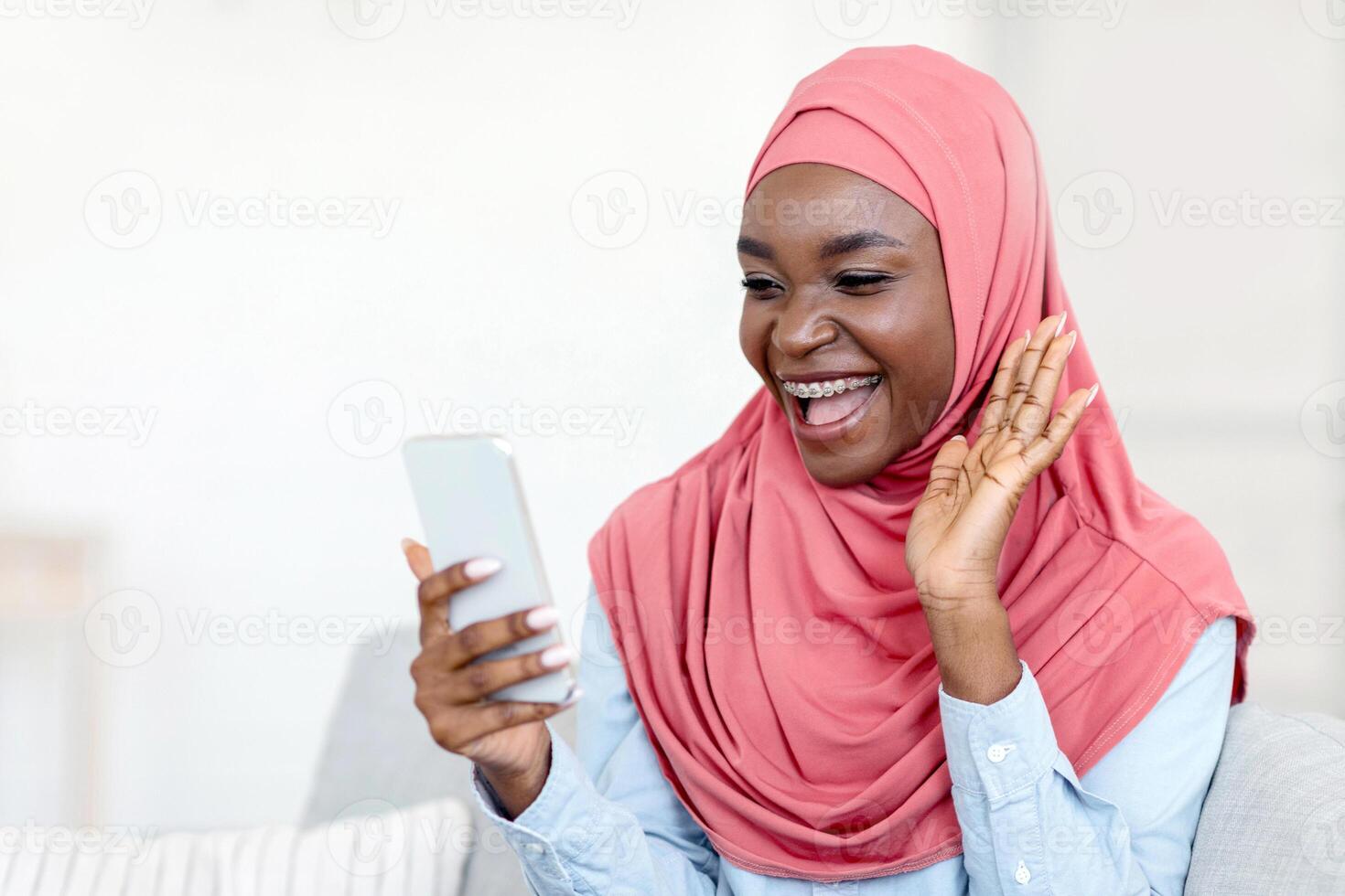 Communication During Quarantine. Black Muslim Woman In Hijab Having Call On Smartphone At Home, Waving Hand At Camera, Talking With Friends While Relaxing On Couch In Living Room, Copy Space photo