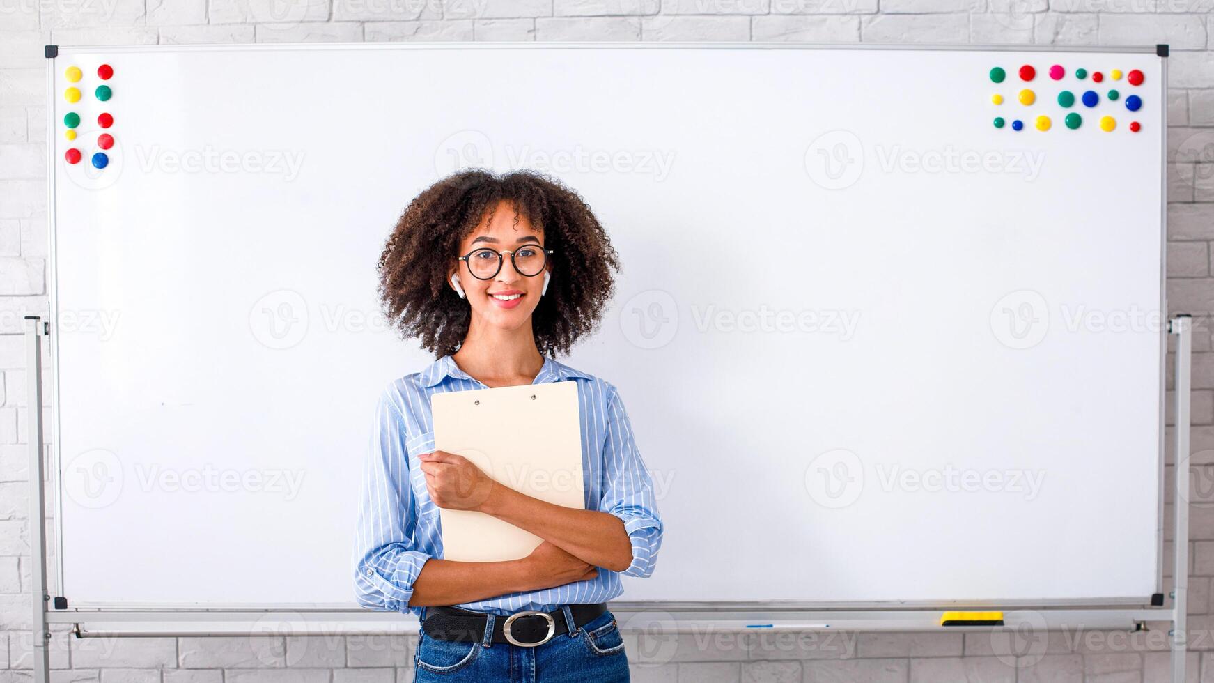 Online work during quarantine and self-isolation. Cute african american girl in wireless headphones and glasses holding notepad on whiteboard background, copy space photo