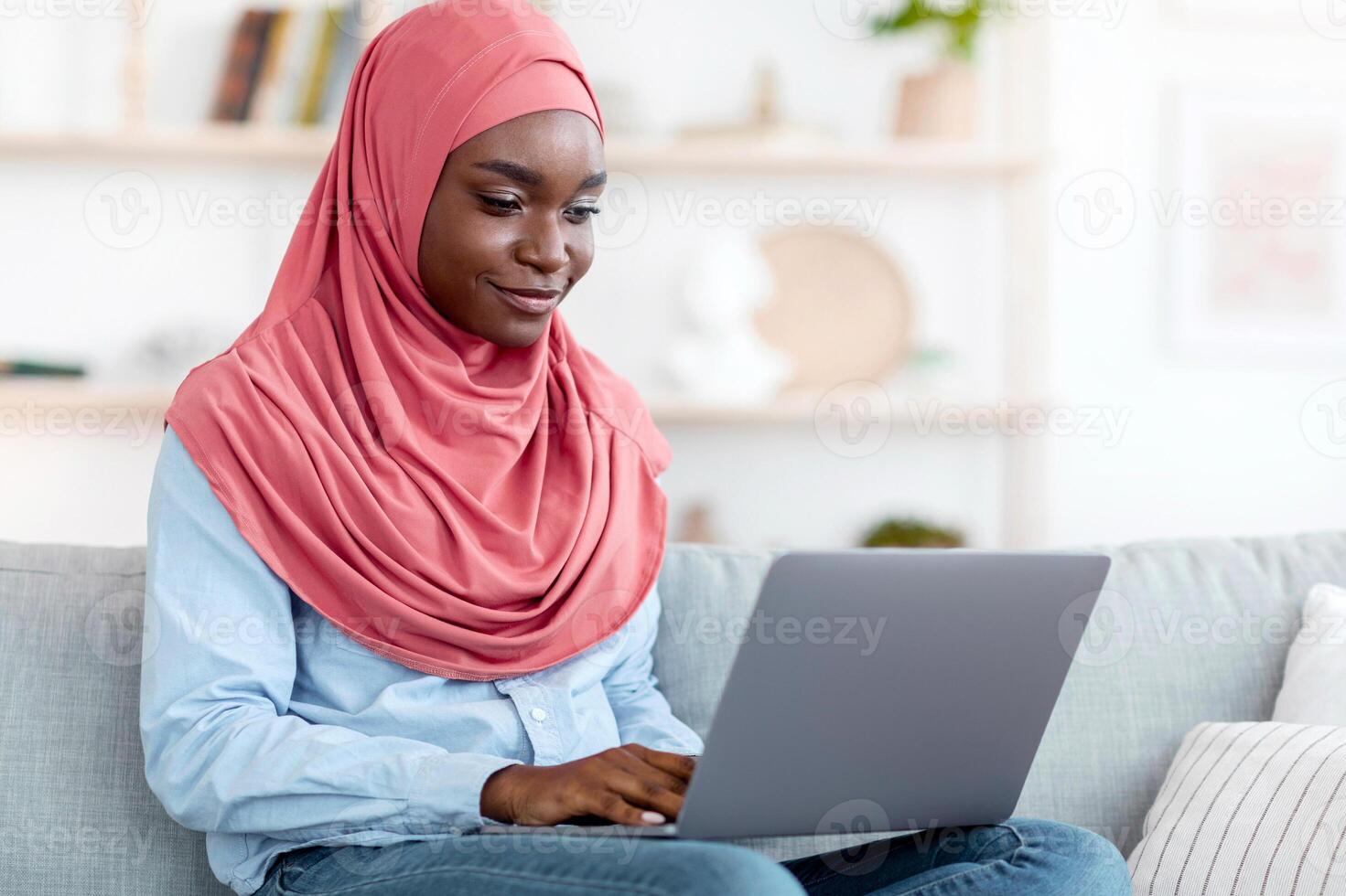 Online Communication. Smiling Black Muslim Woman In Headscarf Using Laptop At Home, African Islamic Lady Messaging With Friends, Typing On Keyboard While Relaxing On Couch In Living Room, Copy Space photo