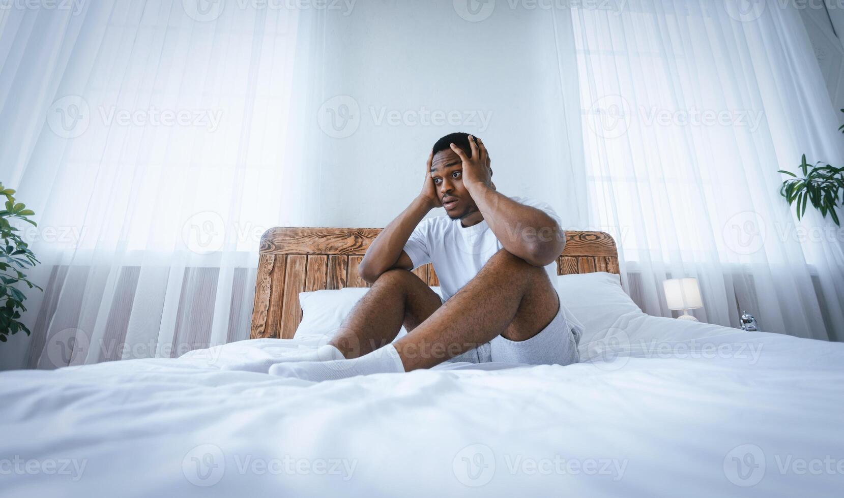 Anxiety And Insomnia. Stressed Black Millennial Guy Sitting In Bed, Touching Head Thinking Of Problems At Home. Panorama, Low-Angle photo