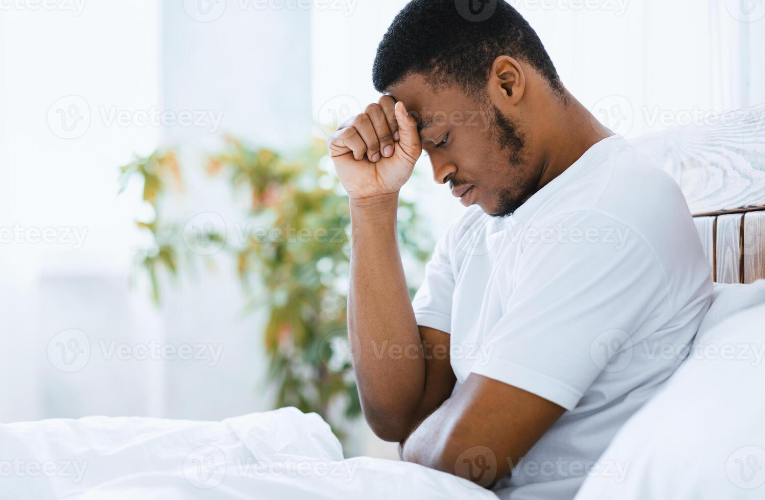 Depressed African American Man Suffering Depression Sitting In Bed Thinking About Problems In Modern Bedroom At Home. Male Mental Health Issue Concept. Free Space For Text photo