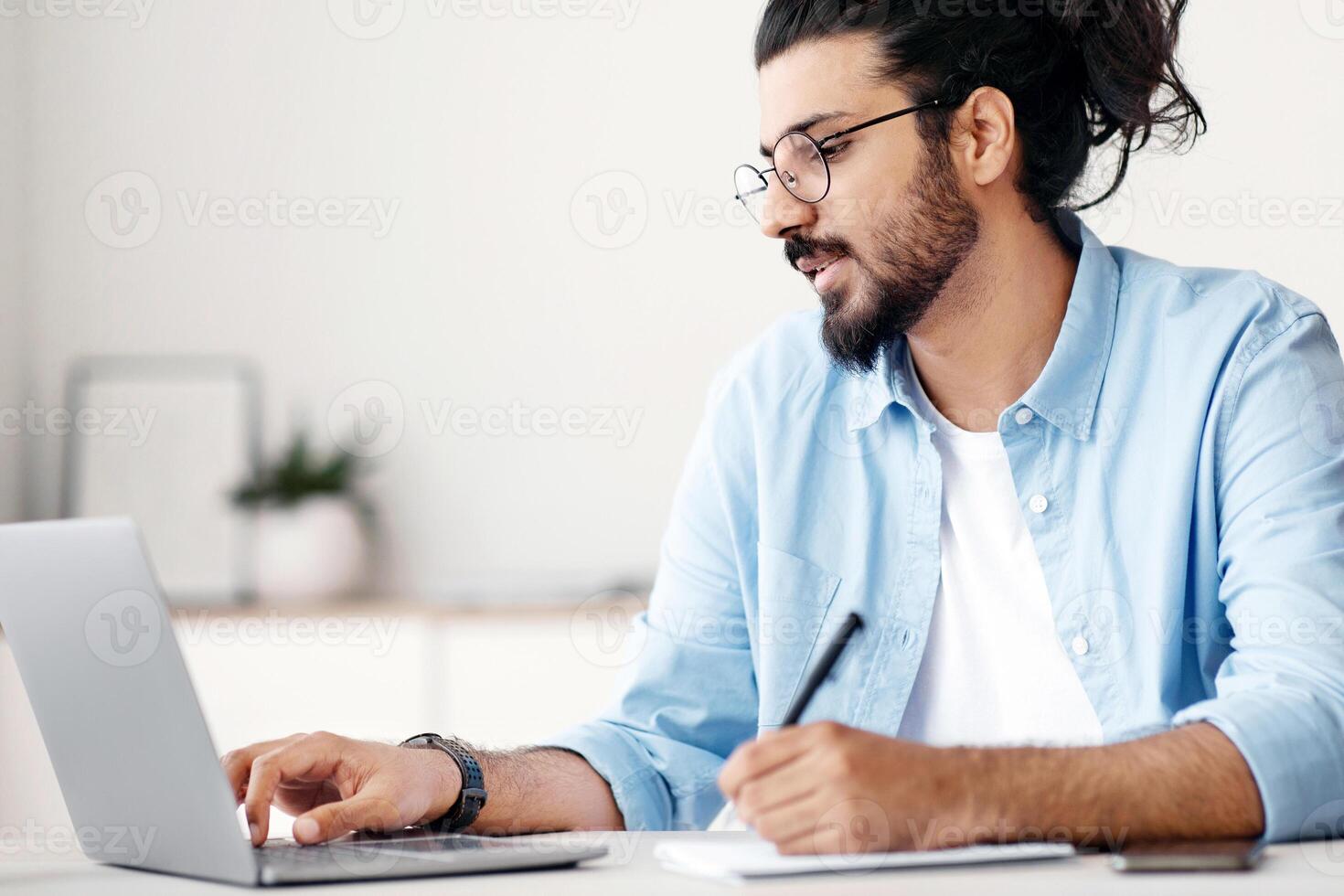Young indian man entrepreneur using laptop and taking notes while working in modern office, sitting at desk and looking at computer screen, wester freelancer guy writing in notepad, copy space photo