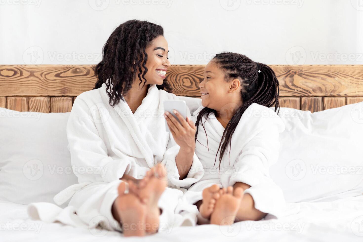 Joyful Black Mom And Daughter In Bathrobes Relaxing With Smartphone In Bed, Browsing Internet, Ordering Food Or Using Cellphone For Online Shopping, Cheerful Mother And Little Girl Having Fun Together photo
