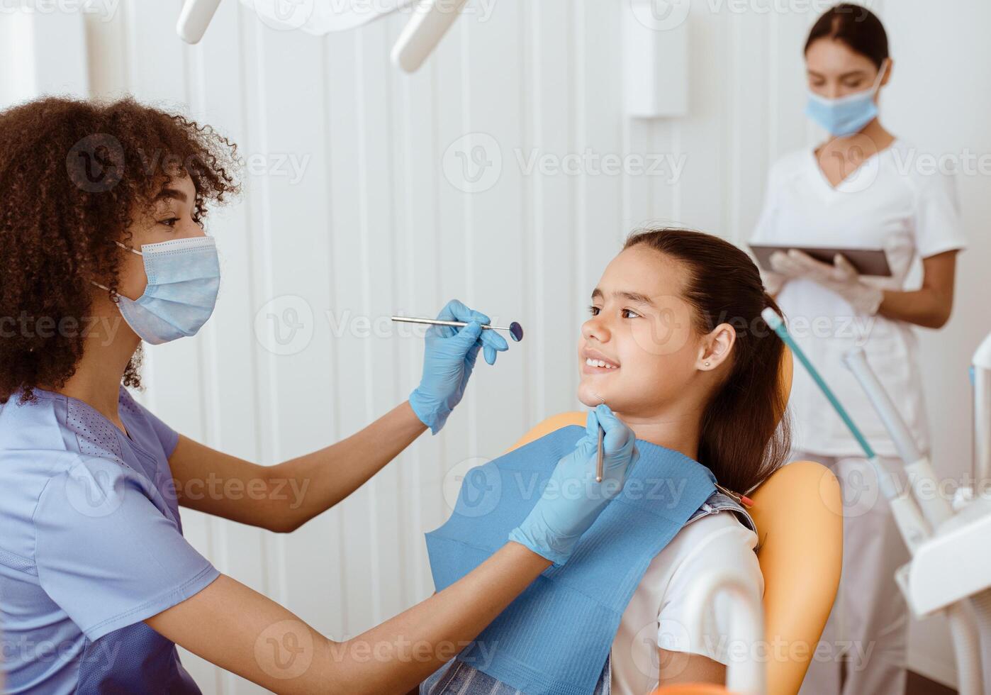 African american dentist and teeth treatment. Happy young doctor and little girl communicate, portrait of stomatologist with team working in background in modern clinic interior, copy space photo