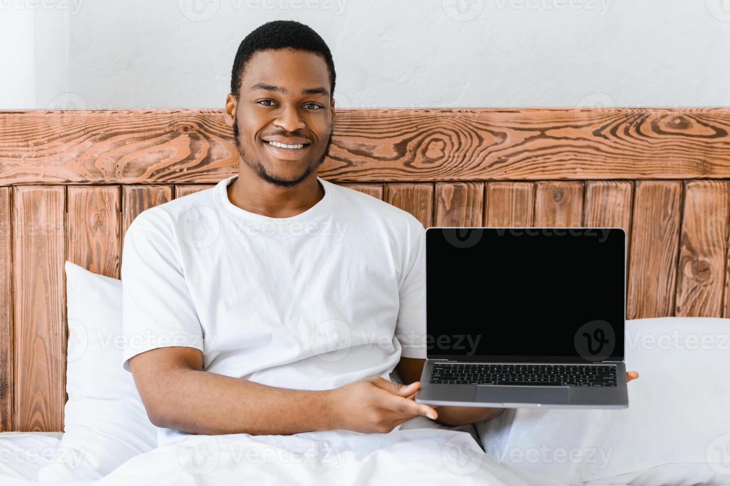 Happy African American Guy Showing Laptop With Empty Screen Advertising Internet Website Sitting In Bed At Home, Smiling To Camera. Computer User Advertising Internet Service Or Web Content. Mockup photo