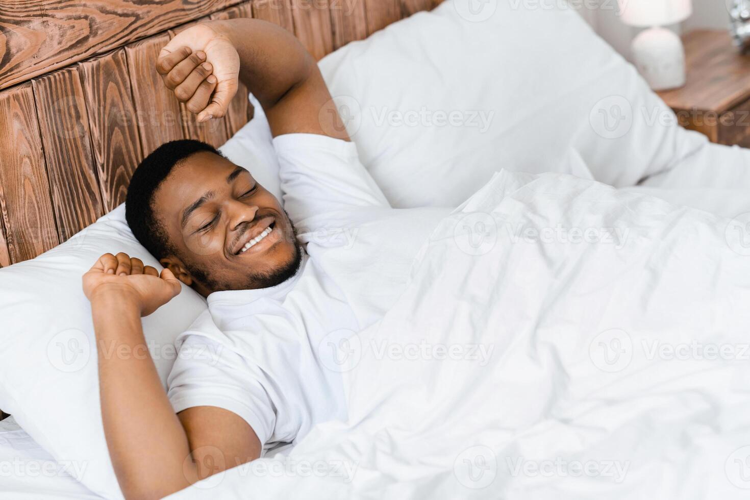 Happy Rested Wellslept Black Man Waking Up In The Morning Stretching Hands Lying In Comfortable Bed In Bedroom At Home, Smiling With Eyes Closed. Good Morning, Awakening Concept. High-Angle View photo