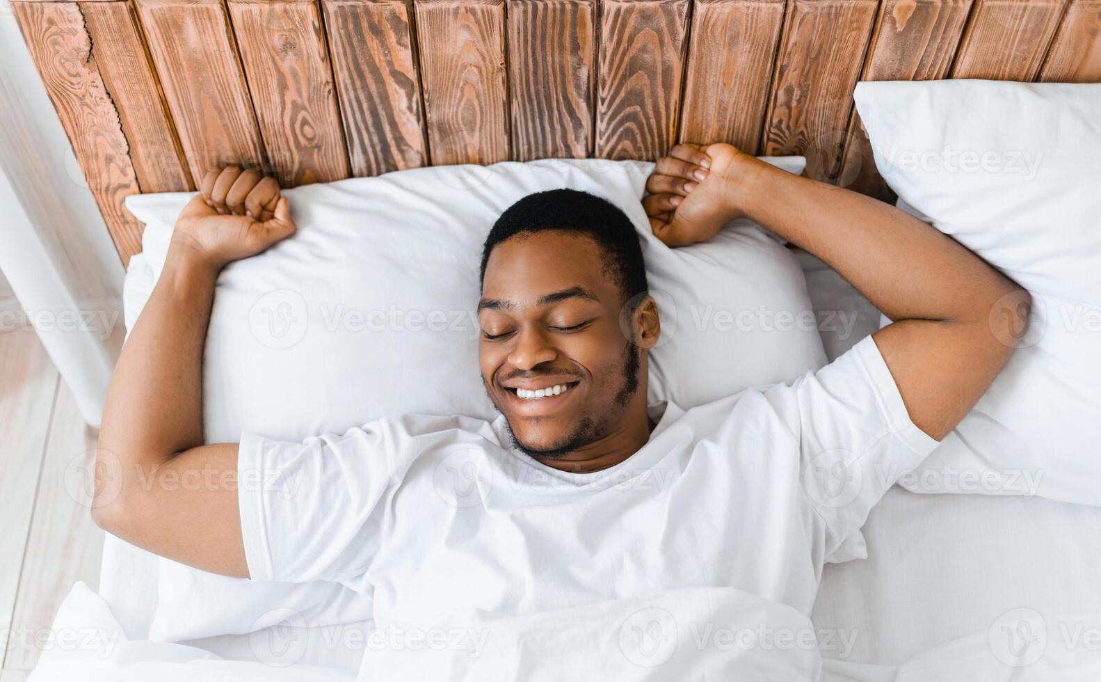 Rested African American Man Waking Up Stretching Hands Awakening Lying In Bed At Home In The Morning, Smiling With Eyes Closed. Good Sleep, Rest And Recreation Concept. Above View photo