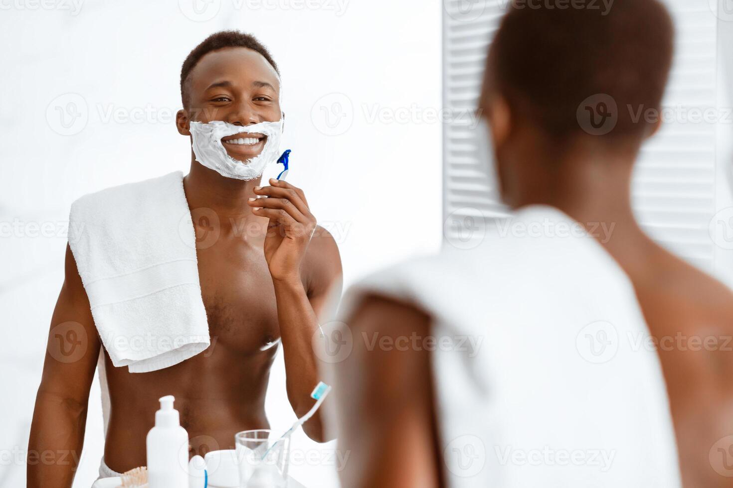 Shirtless African Guy Shaving Face Using Shave Foam And Razor Standing Near Mirror In Modern Bathroom Indoors, Smiling To His Reflection. Male Selfcare Morning Routine And Facial Skincare photo