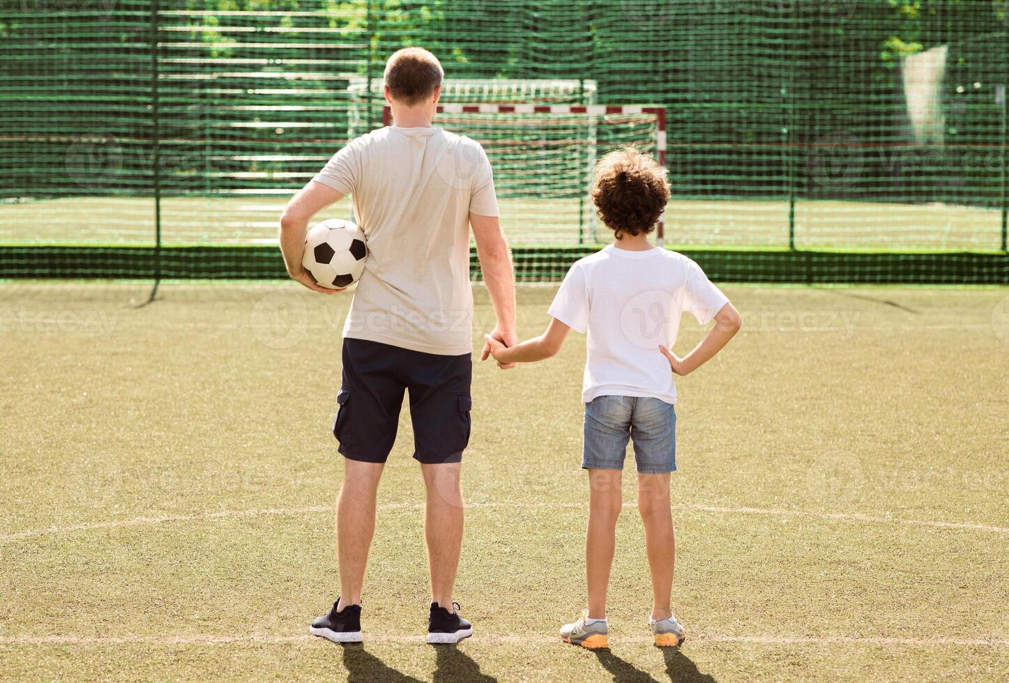 Sports Concept. Back view of dad and son standing at football pitch and looking at goal net, holding hands photo
