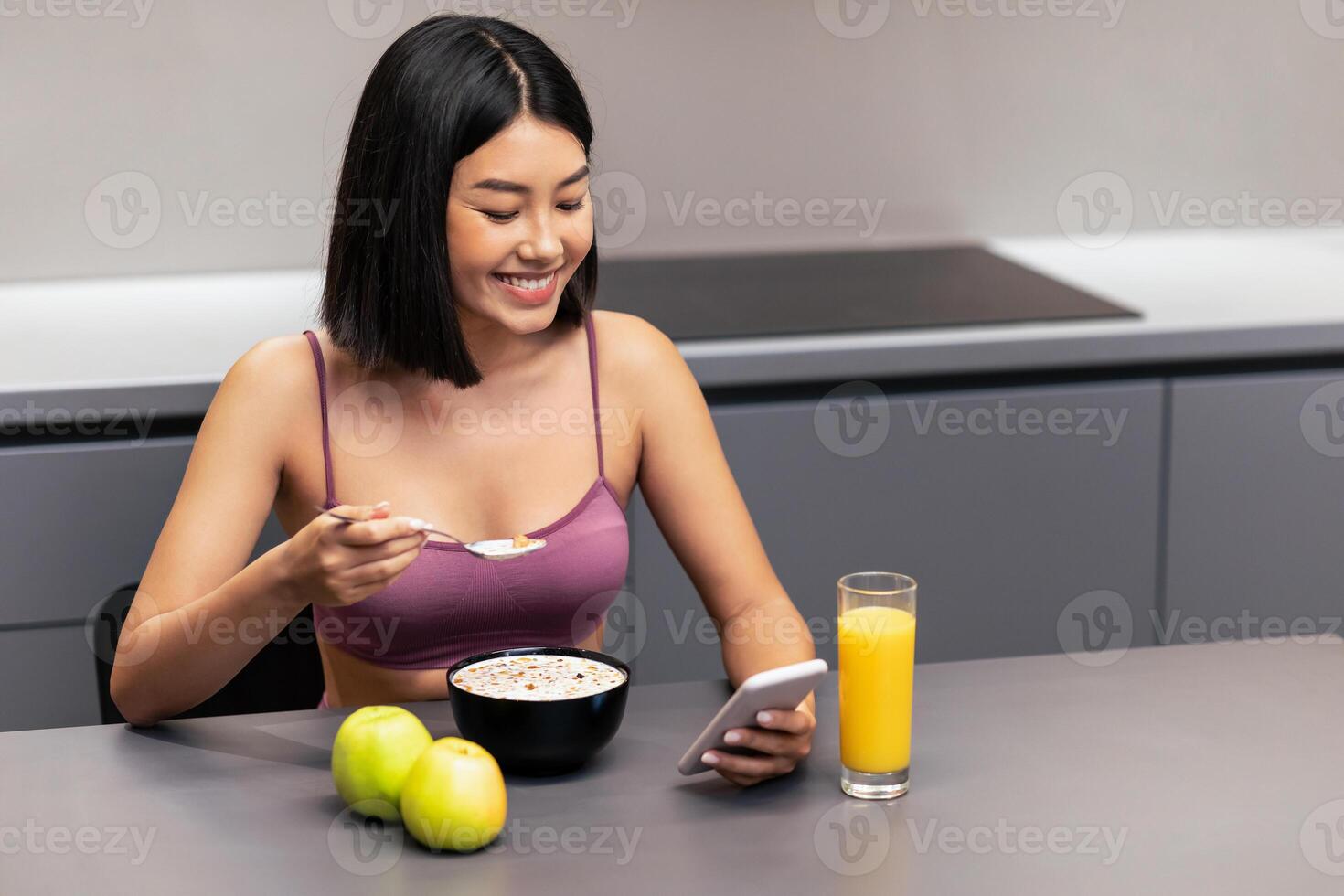 Weight Loss App. Fit Asian Lady Using Smartphone Eating Oatmeal Porridge For Breakfast Sitting At Table In Kitchen At Home. Healthy Diet, Nutrition For Slimming Concept. Empty Space photo
