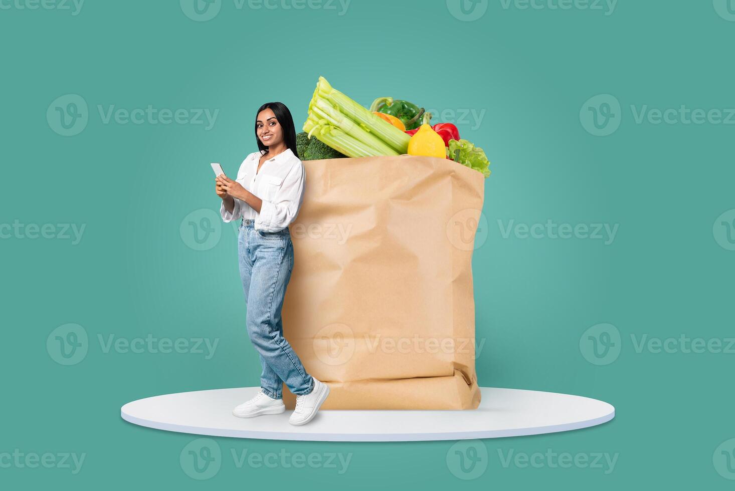 A customer stands next to a large paper grocery bag filled with fresh vegetables and fruits, smiling as she checks her phone for delivery options. This represents modern shopping habits. photo