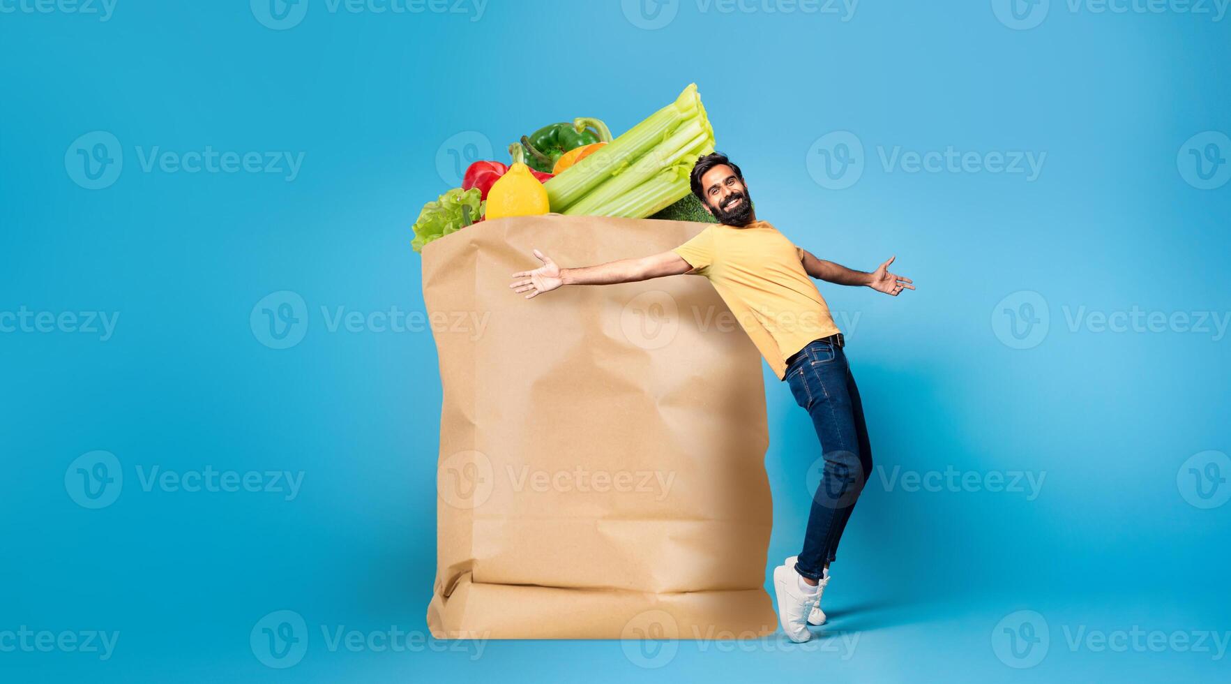 A man in casual attire appears delighted while holding a large paper bag filled with fresh groceries. He stands against a blue backdrop, showcasing the joy of shopping in today's world. photo