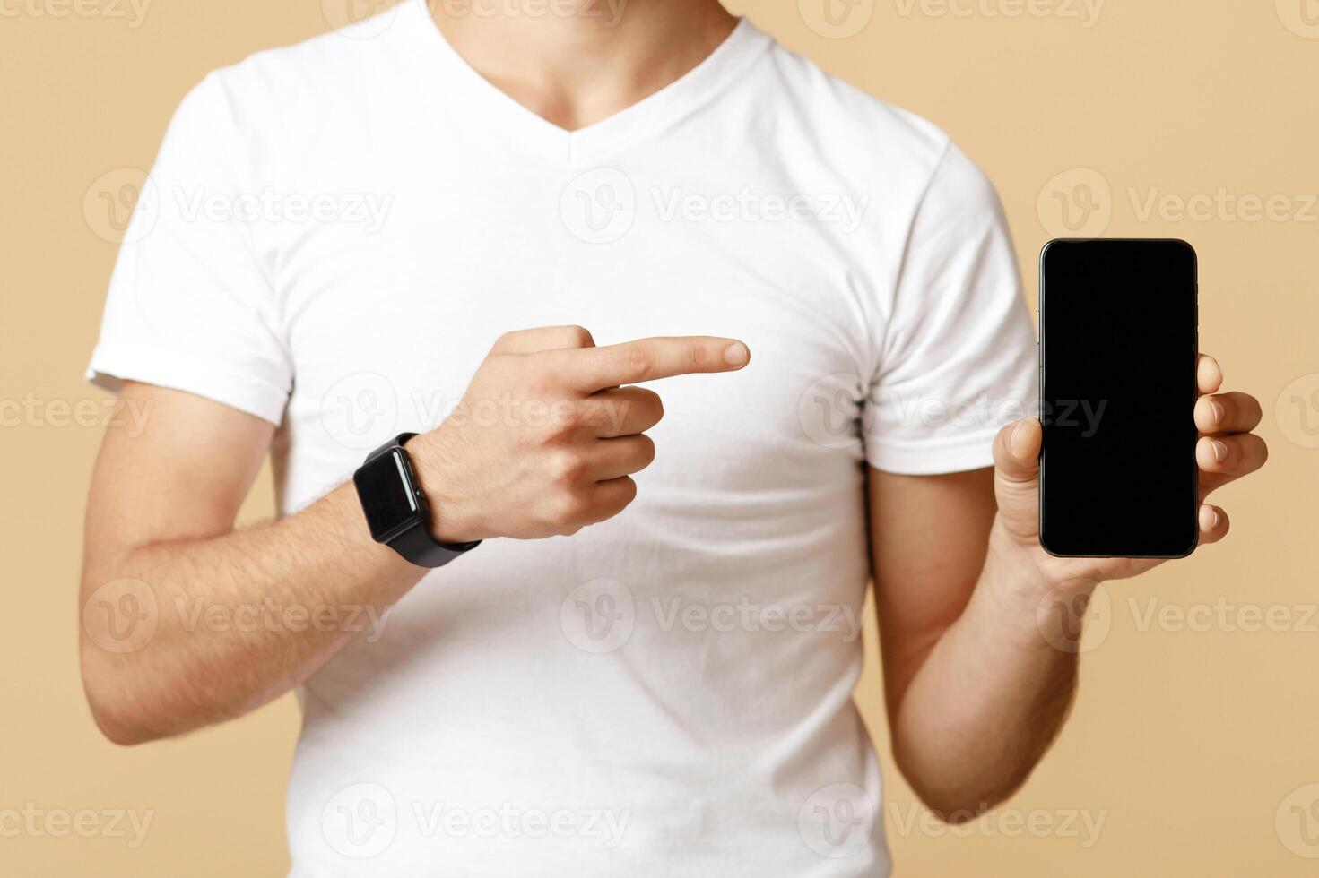 Cropped of young student man in protective mask with glasses shows finger at smartphone with blank screen, isolated on light sand background, studio shot photo