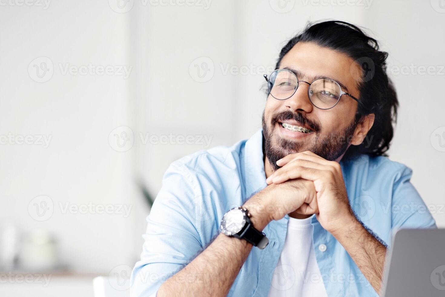 Pensive indian male freelancer thinking about something while working on laptop at home office, sitting at desk and looking away with pleased face expression, wearing glasses and braces, copy space photo