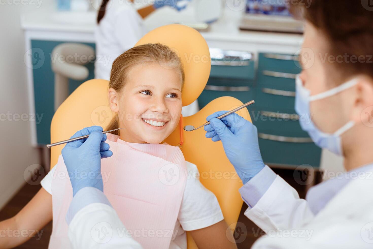 Fearless visit to doctor and dental treatment for child. Smiling girl in medical chair looks at dentist man in white coat, protective mask and rubber gloves with tools in his hands, and not afraid photo