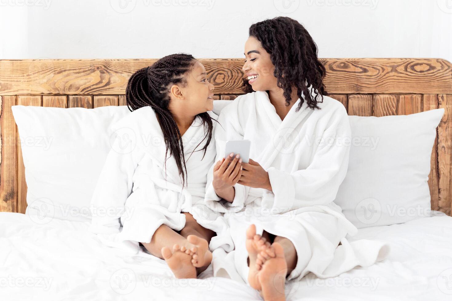 Happy African Mom And Daughter Using Smartphone While Relaxing On Bed In Bathrobes, Browsing Internet, Shopping Online, Checking New App, Resting At Home, Enjoying Time Together, Copy Space photo